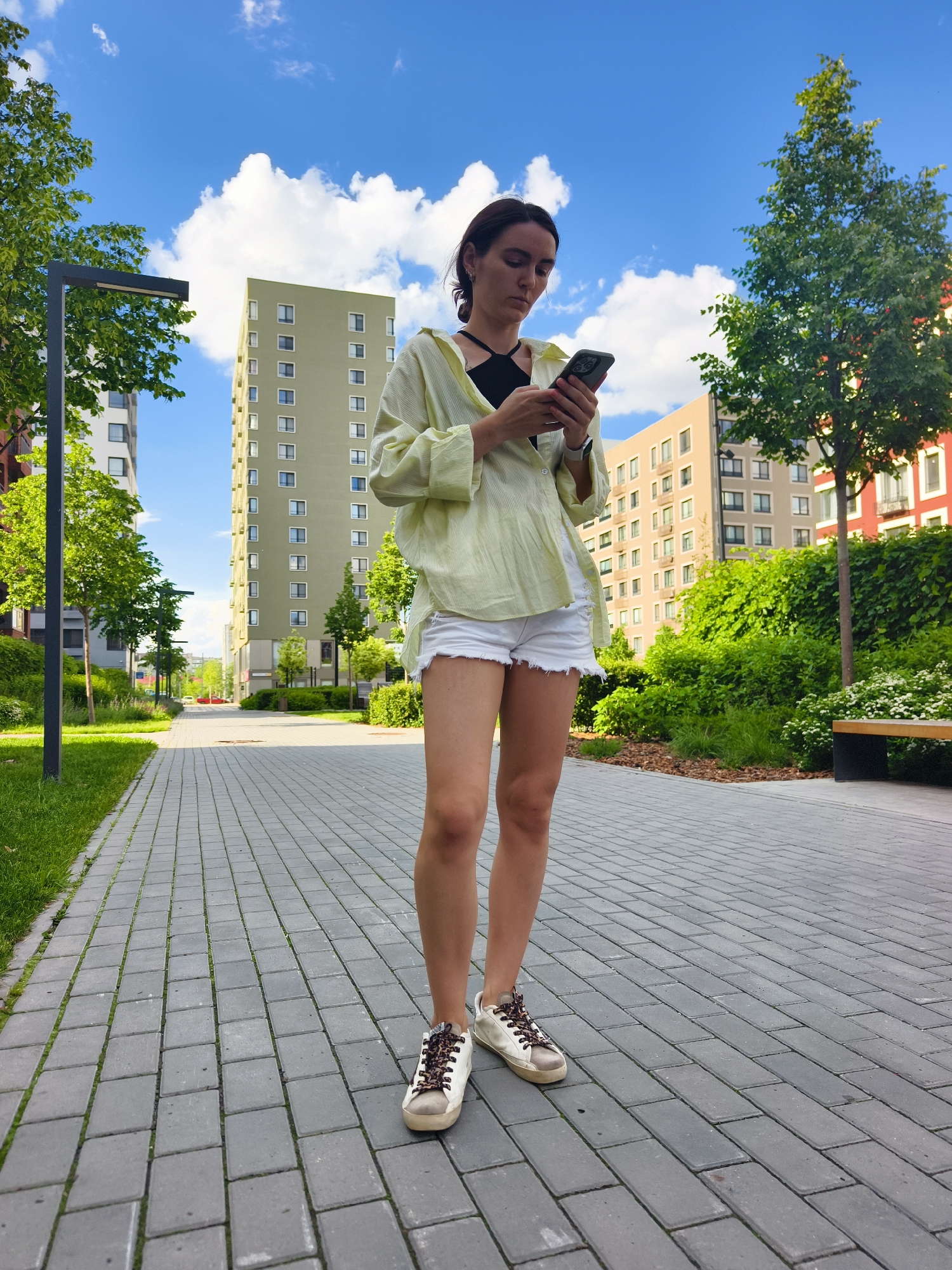 This full-shot image features a young woman standing on a paved walkway in a modern residential complex in Tyumen, Russia, on a bright, sunny day. The camera captures her from a low angle, emphasizing the height of the surrounding buildings and the expansive blue sky dotted with white cumulus clouds.
The woman has medium-length dark hair and is dressed casually in a black top, an unbuttoned, oversized light yellow or lime green collared shirt, white distressed denim shorts, and white sneakers with unique brown and black patterned laces. She is holding a dark-colored smartphone with both hands, her head tilted downwards as she intently focuses on its screen. Her expression is neutral, indicative of her concentration on the device.
The environment is a well-maintained urban landscape. The pathway beneath her feet is constructed from grey rectangular pavers. On either side, there are strips of green grass, various trees, and lush bushes, suggesting a well-landscaped area. In the background, multiple modern apartment buildings with distinct architectural styles and colors—including pale green, beige, and reddish-brown facades—dominate the skyline. A black lamppost is visible on the left, and a contemporary bench with wooden seating and dark metal supports can be seen on the right, nestled among the greenery. The strong natural light and shadows indicate it is likely midday or early afternoon. No discernible text is visible in the image.