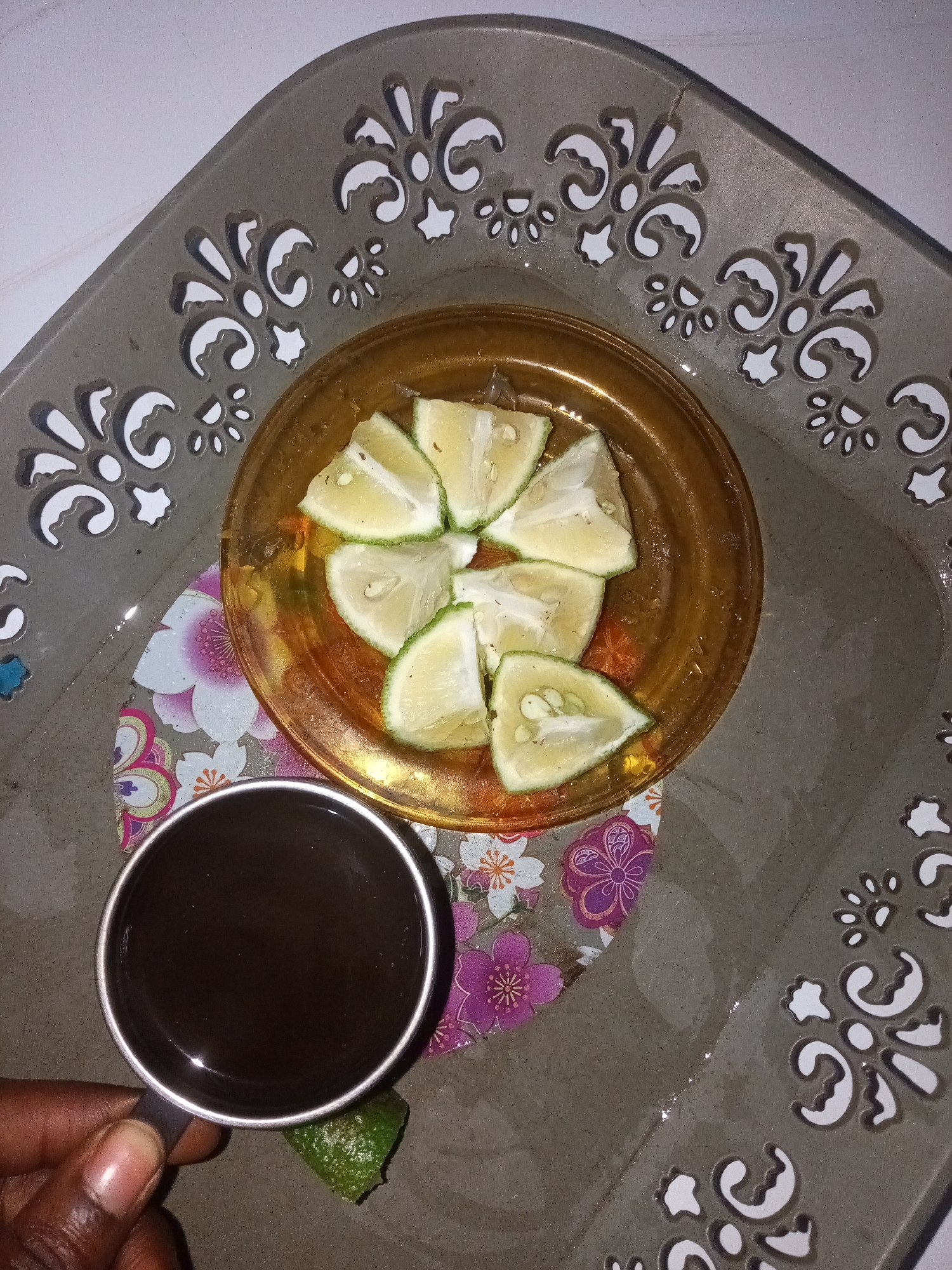 A hand holding a mug of dark liquid is in the bottom left corner of the image. The mug is positioned on a textured gray tray with decorative cutouts along its rim. To the right of the mug, a small piece of green lime is visible. In the center of the tray, a small amber-colored dish holds several slices of lime arranged in a circular pattern. Beneath the amber dish, a small, colorful coaster with floral designs can be seen. The background is a light-colored surface, likely a table. The scene is likely located in New Stadium, Nigeria, given the context.