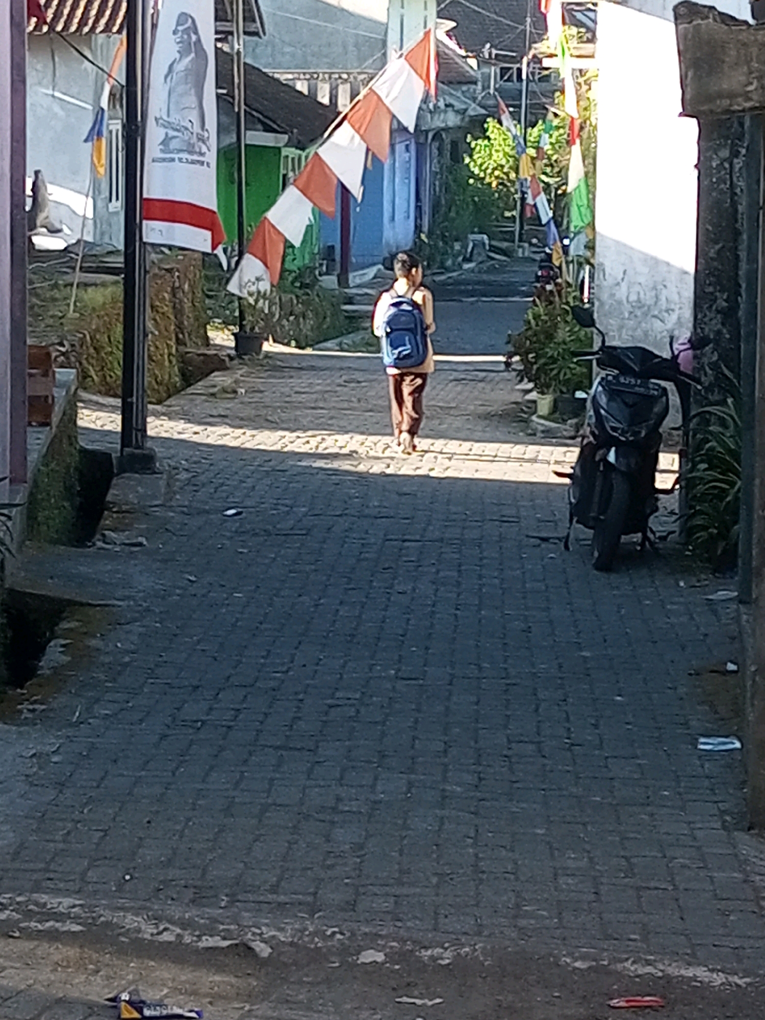 A narrow, paved alleyway in Andongsili, Indonesia, is captured from a low angle looking uphill. The alley is lined with houses and vegetation. In the foreground, a black motorcycle is parked on the right side of the alley. A young person, facing away from the camera, walks up the alley carrying a blue backpack. The alley is decorated with Indonesian flags and other colorful banners. The pavement is made of interlocking bricks. Debris is scattered on the ground along the sides of the alley.