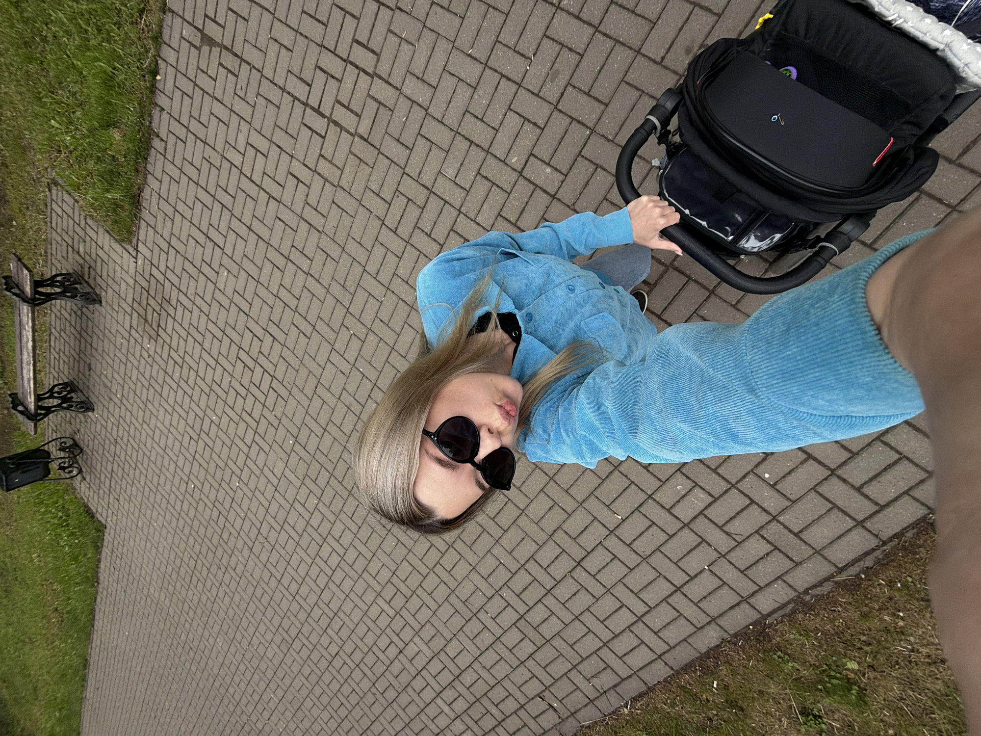 A high-angle selfie captures a person pushing a baby stroller on a paved path outdoors. The subject, likely female, has light-colored hair and wears dark, round sunglasses. She is dressed in a textured, light blue button-up jacket and grey pants, with black shoes peeking out. Her expression features slightly pursed lips. She is holding the handle of a black baby stroller, which has a structured frame and a compartment containing various items, including a small purple object and a metallic pin or clip on a black surface.
The setting is an outdoor area in Chaikovsky, Russia, characterized by a wide path made of grey rectangular paving stones laid in a herringbone pattern. To the left of the path is a strip of green grass, and a dark-colored park bench with ornate metal legs is partially visible further back on the grass. The lighting appears to be natural daylight, possibly under an overcast sky, as no strong shadows are cast. No visible text is present in the image. The scene depicts a casual moment, likely a parent or caregiver out for a walk with a child.