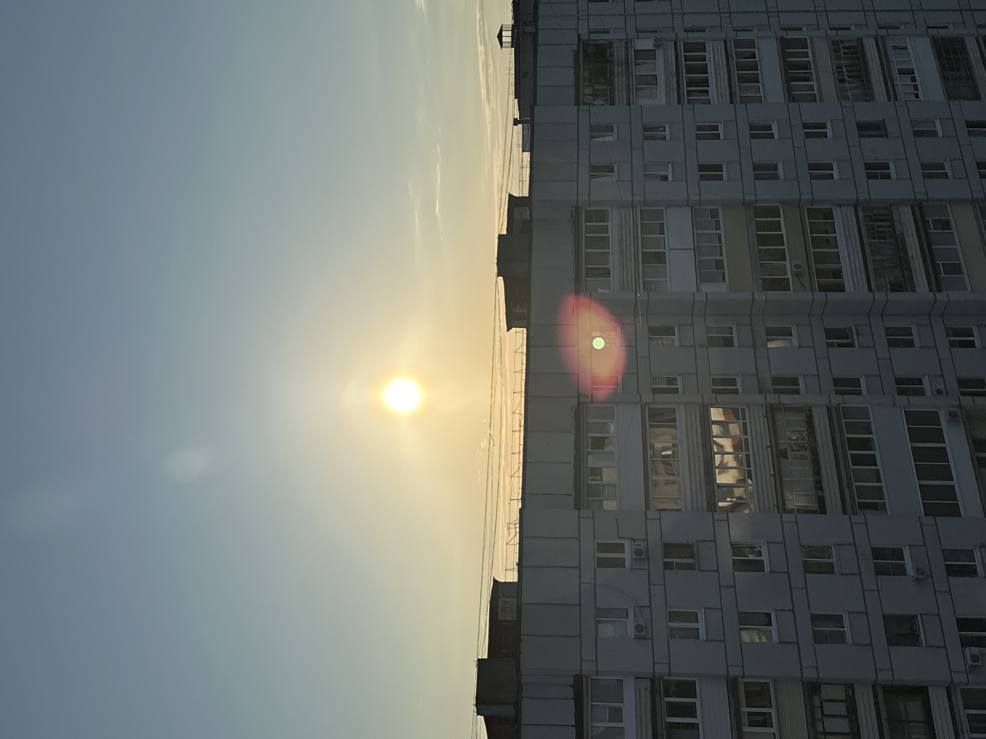 The image shows a high-rise building in Chaikovsky, Russia, at sunset. The sky is light blue near the top and transitions to a yellowish hue near the horizon where the sun is visible as a bright white disk with a lens flare. The building, a tall concrete structure, occupies the right side of the frame, its many windows arranged in a regular grid. Some windows have air conditioning units. The building appears to be under renovation or construction, as some scaffolding is visible on the left side of the building's edge.