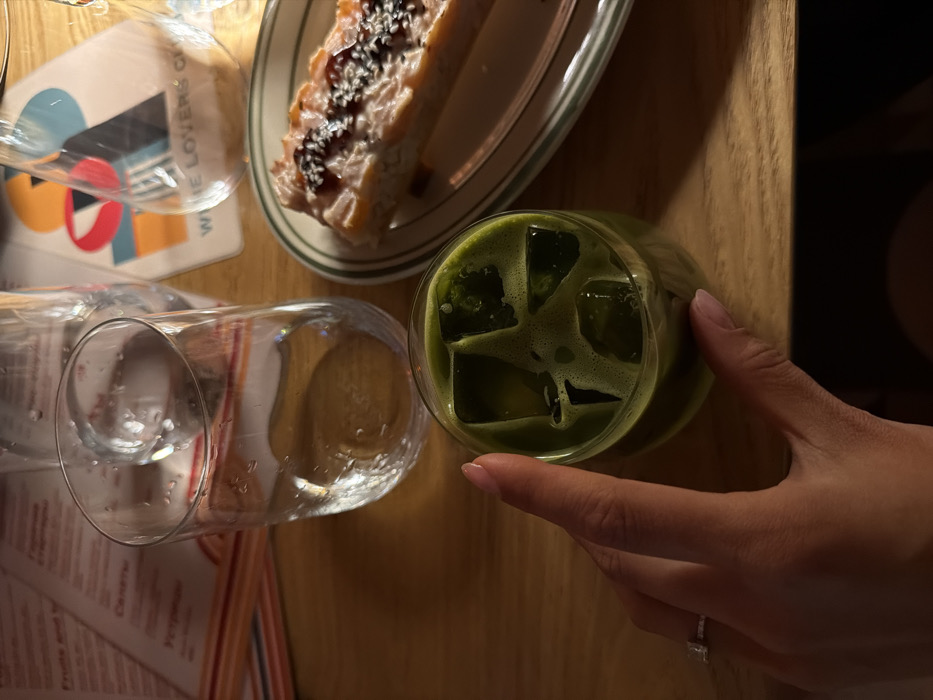 A wooden table displays a glass of iced matcha, a dessert slice on a green-rimmed plate, and a glass on top of a colorful coaster. A hand with a ring delicately holds the matcha glass, hinting at a dining setting. The composition is an overhead shot, showcasing the textures of food and drink alongside the table's surface.