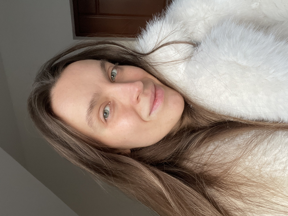 A woman poses in a close-up shot, wearing a soft, white fur garment. She has light brown hair, fair skin, and green eyes. Her expression is relaxed and content. The background is simple, with a partially visible wooden frame at the top and a gray wall on the left. The photo is slightly rotated, giving a casual and intimate feel.