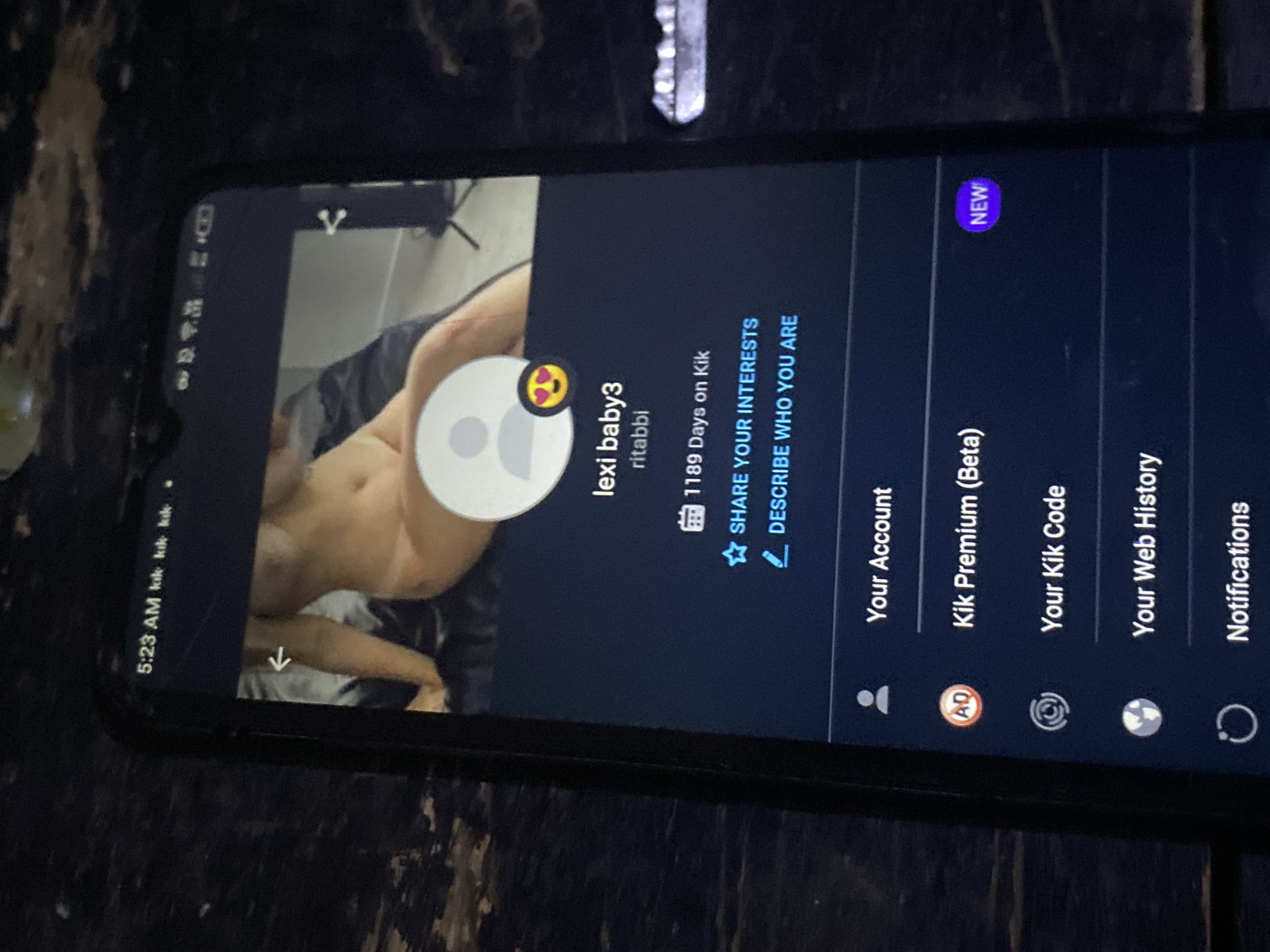 A smartphone displays the Kik messaging application. The screen shows a Kik profile of a user named "lexi baby3" with the username "ritabbi". A semi-nude person is in the background of the profile picture. The menu options displayed include "Your Account", "Kik Premium (Beta)", "Your Kik Code", "Your Web History", and "Notifications". The phone's status bar shows the time as 5:23 AM. The Kik notification icons are visible. The phone is lying on a dark, wooden surface. The image does not contain any geographical information that confirms this image originated in Bukuru, Nigeria.