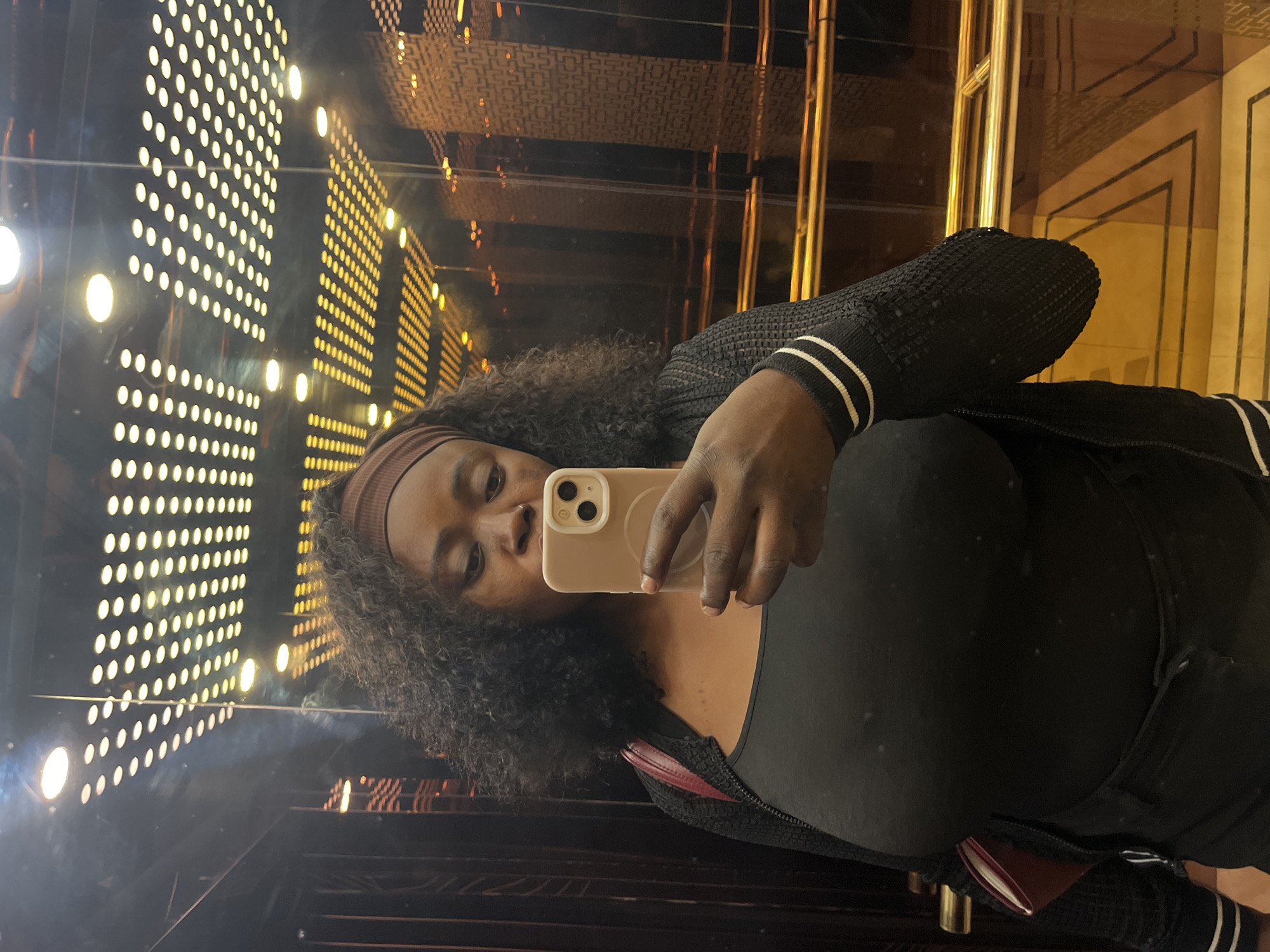 A medium shot captures a Black woman taking a mirror selfie in an elevator. She is wearing a black tank top, a black zip-up jacket with white stripes on the cuffs, black pants, and a brown headband. Her dark, curly hair frames her face as she holds a light pink smartphone in her right hand. The elevator's mirrored walls reflect the woman and the surroundings. The ceiling is illuminated by rows of small, circular lights arranged in a grid pattern. The elevator walls are paneled with dark wood and feature gold railings. The floor has a geometric pattern with gold accents. The overall impression is that of a modern and well-lit elevator. The image does not contain any specific visual cues to pinpoint its location within Abuja, Nigeria.