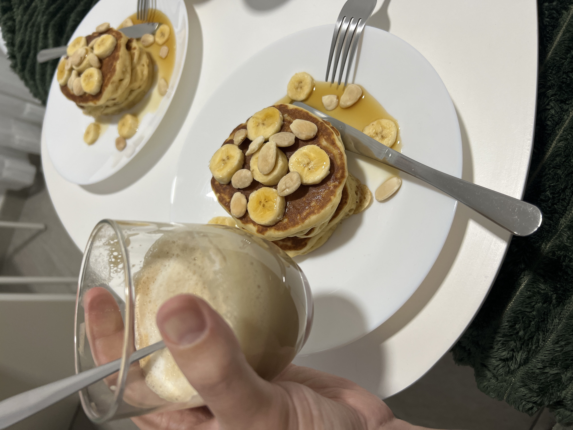 The image shows a tabletop setting with two plates of pancakes, a drink, and cutlery. Each plate holds a stack of pancakes topped with sliced bananas and almonds, drizzled with syrup. A fork and knife rest on each plate. In the foreground, a hand holds a glass containing a foamy beverage, likely coffee or a latte, with a spoon inside. The background suggests an interior setting with white furniture and a hint of a green, textured fabric. No evidence suggests this photo was taken in Mogán, Spain.