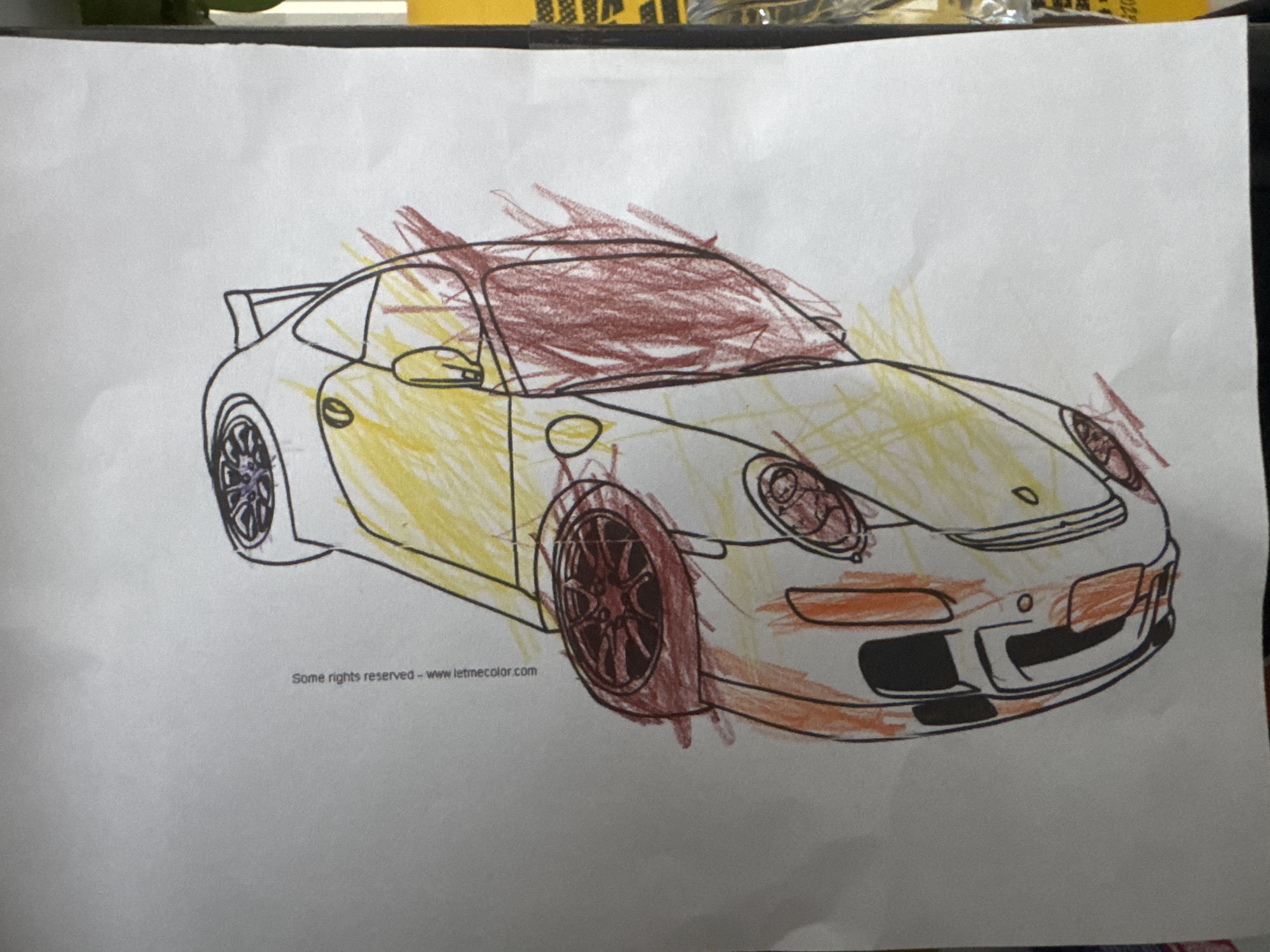 The image shows a crayon drawing of a Porsche 911 on a white sheet of paper. The car is mostly yellow with red crayon shading. The location is unknown.