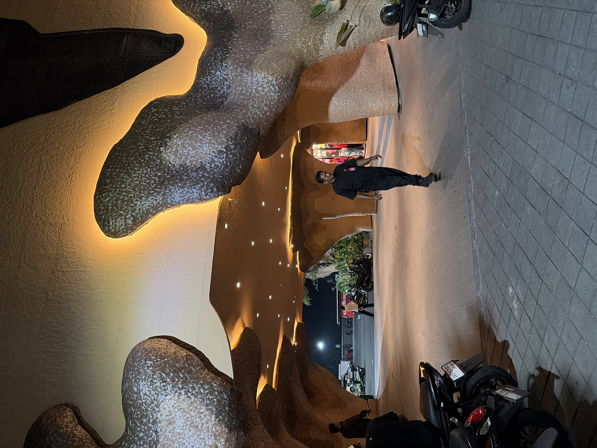 In a dimly lit outdoor setting, possibly a walkway or entrance to a establishment, a person dressed in a black uniform stands facing away from the camera. The person appears to be a security guard, given their attire. Behind them, the architecture features an undulating, organic design with textured surfaces in shades of beige and grey, illuminated by warm, indirect lighting. This creates a cave-like or naturalistic ambiance. Small, circular lights are embedded in the ceiling further down the walkway.
To the right of the walkway, a tiled surface suggests a paved area. Several motorcycles are parked, with one partially visible in the foreground. The time of day appears to be evening or night, given the artificial lighting and overall darkness. The general atmosphere is quiet and somewhat mysterious due to the lighting and the unusual architectural elements. No specific weather conditions are discernible.
The scene is set in Ubud District, Indonesia, which provides a cultural and geographical context for the image, suggesting a tropical environment possibly within a resort, restaurant, or entertainment venue. The image is oriented vertically, with the main subject and architectural features appearing on the left and right sides respectively when viewed in portrait mode.