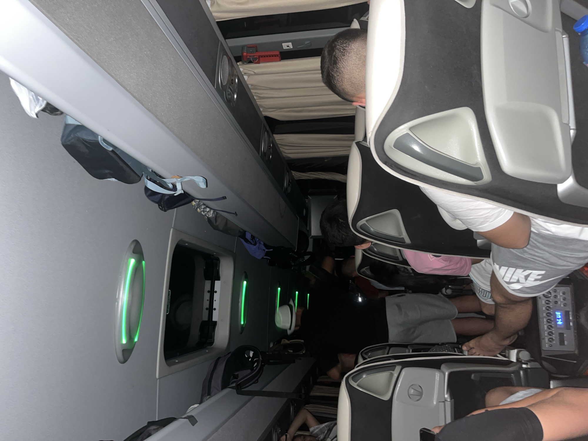 The image depicts the interior of a bus, likely a passenger bus, with seats arranged in rows. Passengers are seated, with some appearing to be in a relaxed or sleeping posture. Overhead storage compartments are visible above the seats, holding various bags and personal items. Cabin lights, possibly LED, are illuminated with a green glow, accentuating the windows along the side of the bus. The timestamp "21:34" can be seen, indicating the time of day. There is no information in the image that directly associates the location with the city of Vinogradets, Bulgaria.