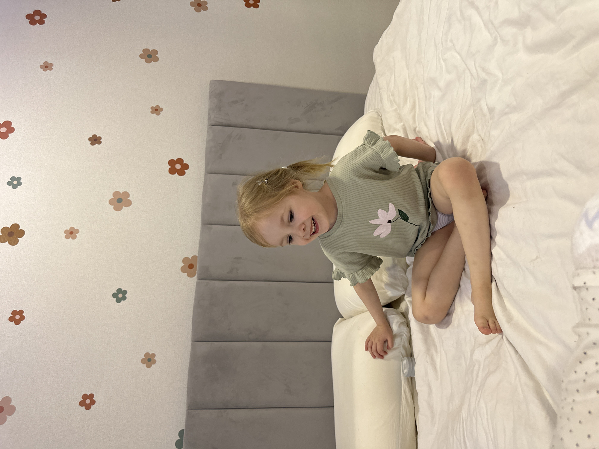 The image displays a fair-skinned child, estimated to be between two and four years old, with light blonde hair styled in two small pigtails secured with clips. The child is seated cross-legged on a bed covered with white bedding. They are wearing a light green, short-sleeved top with ruffled sleeves and a pink flower graphic on the front, over white underwear. The child is smiling, looking towards the viewer's left, with their mouth open, revealing teeth. Their right hand rests on a white pillow positioned to their left. A small, round, grey container with a translucent lid is situated on the bedsheet near the pillow. Behind the child and pillows is a grey, upholstered headboard composed of vertical panels. The wall above the headboard is light-colored and decorated with numerous scattered flower decals in shades of brown, pink, and teal. The specific location within the city of Chaikovsky, Russia, cannot be determined from the visual information provided in the image; the scene depicts an indoor domestic bedroom setting.