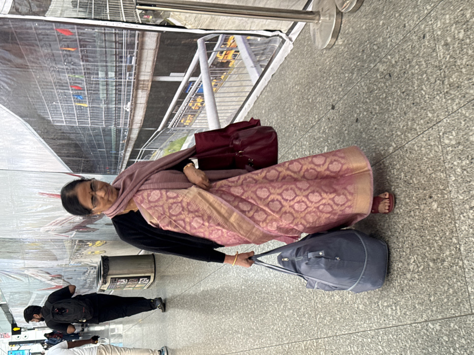 An older Indian woman in a patterned pink sari, black sweater, and glasses pulls a gray duffel bag in an airport or similar indoor space. She carries a burgundy purse. A man in black clothes and a backpack walks nearby. In the background, a large banner displays a stadium or transportation hub scene. The floor is tiled.
