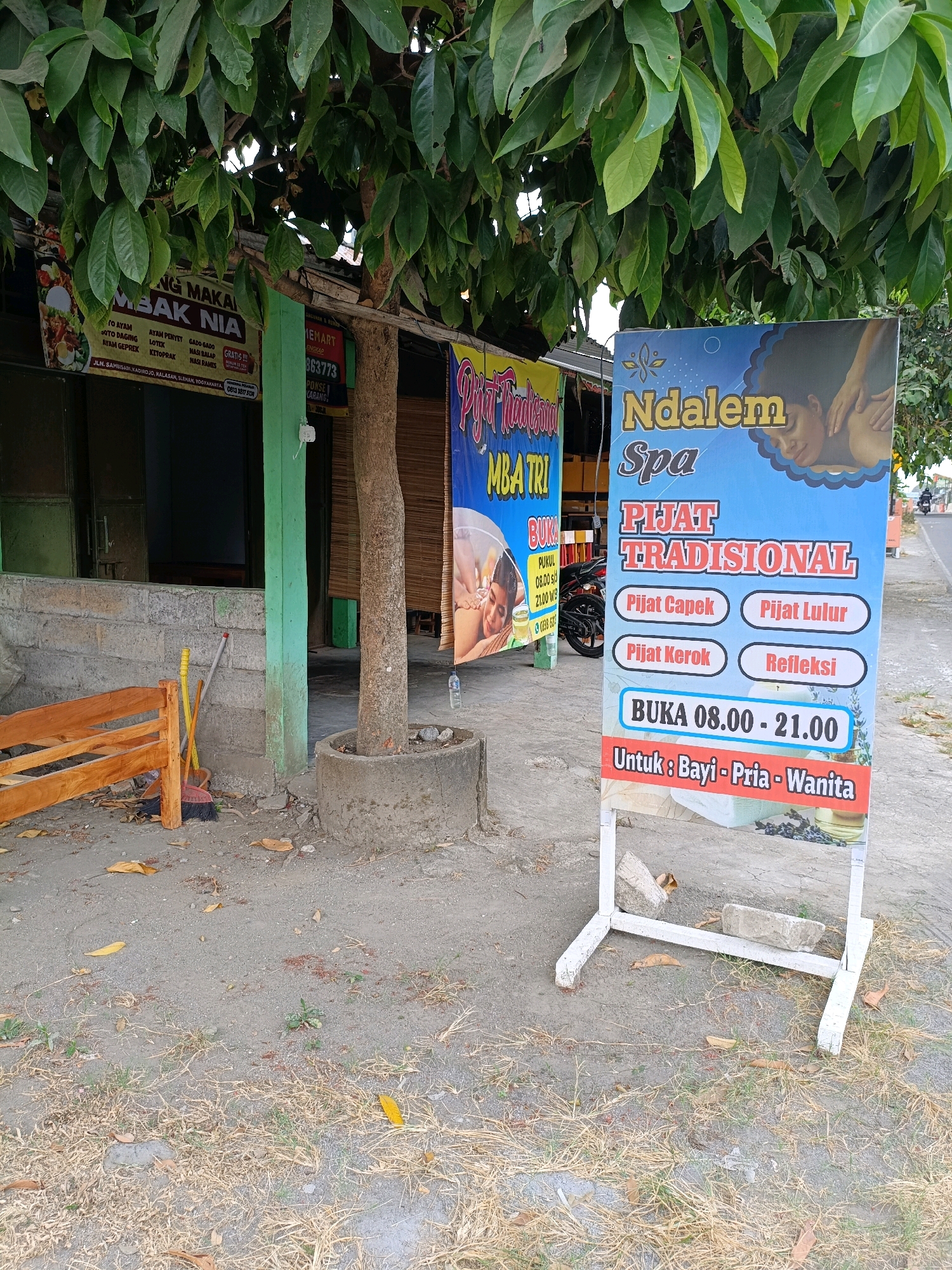 The image shows a street-side view of businesses in Purwomartani, Indonesia. In the foreground, a white A-frame sign displays advertising for "Ndalem Spa" offering "Pijat Tradisional". The sign lists services such as "Pijat Capek", "Pijat Lulur", "Pijat Kerok", and "Refleksi". It also indicates operating hours are from 08.00 to 21.00 and that the spa is for "Bayi - Pria - Wanita". To the left of the spa sign, a wooden bench and cleaning tools including a broom and dustpan are visible. Behind these, a building with a green painted pillar and dark windows is situated. Further to the left, another banner advertises "MAKMBA NIA", listing various food items. The scene is set under the canopy of a leafy tree, with dry leaves and sparse grass on the ground. A road with a motorcycle passing by is visible in the background on the right.