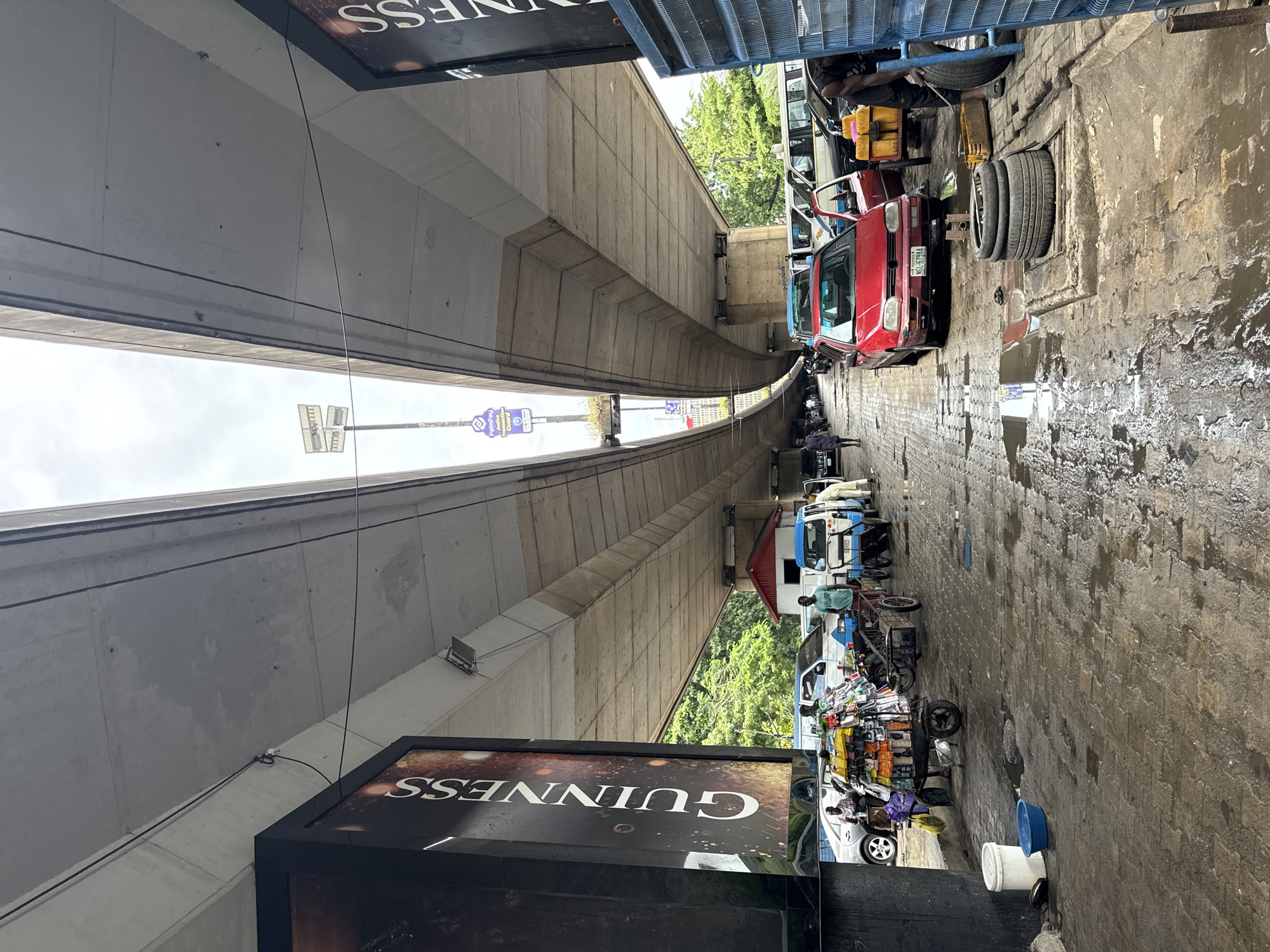 This is a ground-level, eye-level, medium shot of a busy street under an elevated highway in Ikoyi, Nigeria. The concrete structure of the highway dominates the upper portion of the frame, with its underside casting shadows on the scene below. A large, vertical Guinness advertisement is prominently displayed on the left, with text running vertically.
The street itself is paved with interlocking bricks and is wet and reflective, indicating recent rain or a street cleaning operation. Several vehicles are present, including a red car parked in the foreground with its door open, a blue and white minibus, and other cars and vans further down the street. There are people on foot, some appearing to be vendors with carts piled high with goods, and others walking or tending to vehicles.
On the right side of the frame, a man is shown working on tires, with a stack of tires next to him and a yellow container. The environment appears to be a commercial area with a mix of vehicular traffic and pedestrian activity. The overall impression is one of a lively, urban street scene.