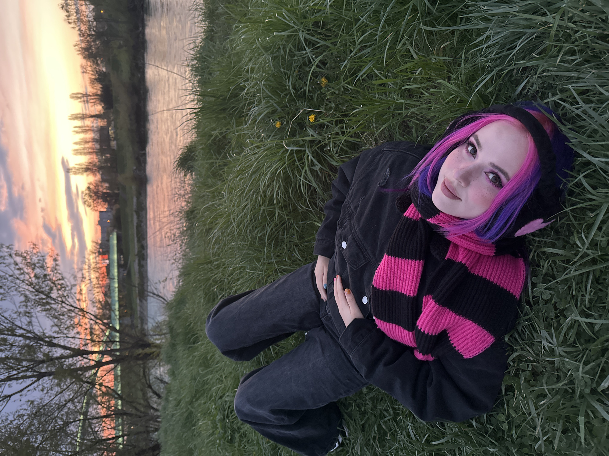 The image captures a scene outdoors, possibly in a park near a river or body of water, judging by the presence of lush green grass and a landscape mirroring the colors of the sunset in the water. The sky shows shades of pink and yellow as the sun sets, painting the clouds and the surrounding trees in a warm glow. A person is lying on the grass, facing upwards. They are wearing dark-colored clothing, consisting of a jacket and jeans, and have a distinctive pink and purple-colored hair with black and pink accessories or a hooded scarf around their head. The scene is suggestive of a relaxed setting, possibly along the banks of the Sava river in Zagreb, Croatia. However, without specific landmark information, a precise location within Zagreb cannot be definitively determined.