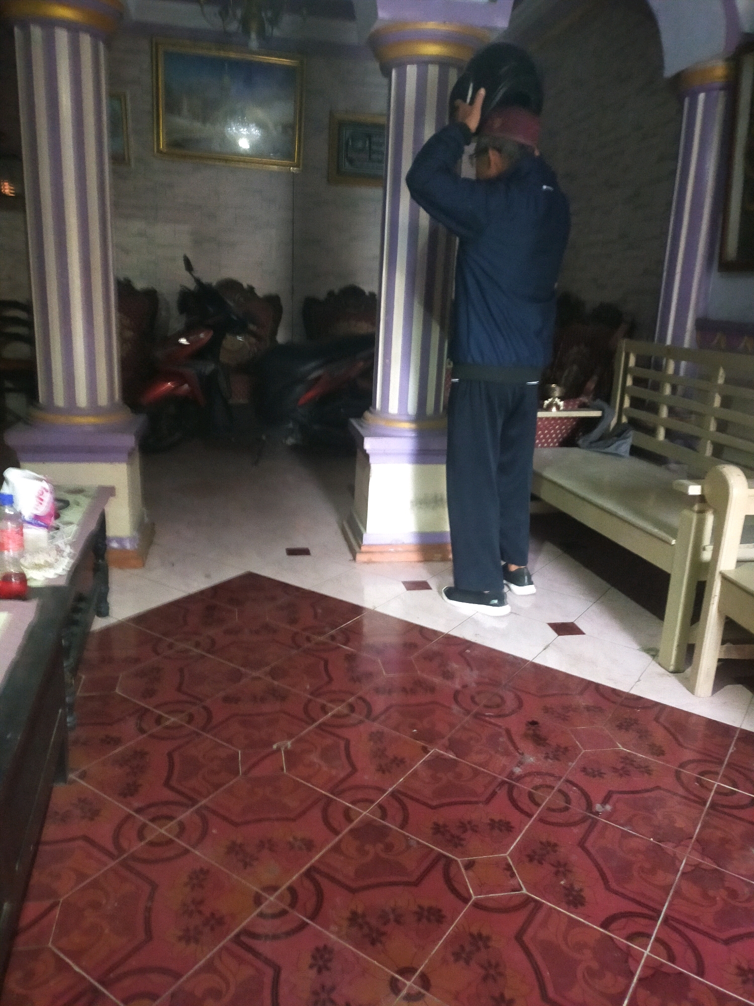 A person wearing a dark blue jacket, black pants, a maroon scarf, and a black helmet is standing in what appears to be the entryway of a home in Mojodanu, Indonesia. The person is in the process of putting on or adjusting their helmet. The floor is covered in red patterned tiles. In the background, there are two motorcycles parked, wooden furniture, and large decorative pillars with purple and white stripes. On a table to the left, there are a few items including a bottle of water and a packet of tissues. The walls are decorated with framed artwork. A wooden bench is visible to the right of the person. The lighting suggests it is either early morning or late evening.