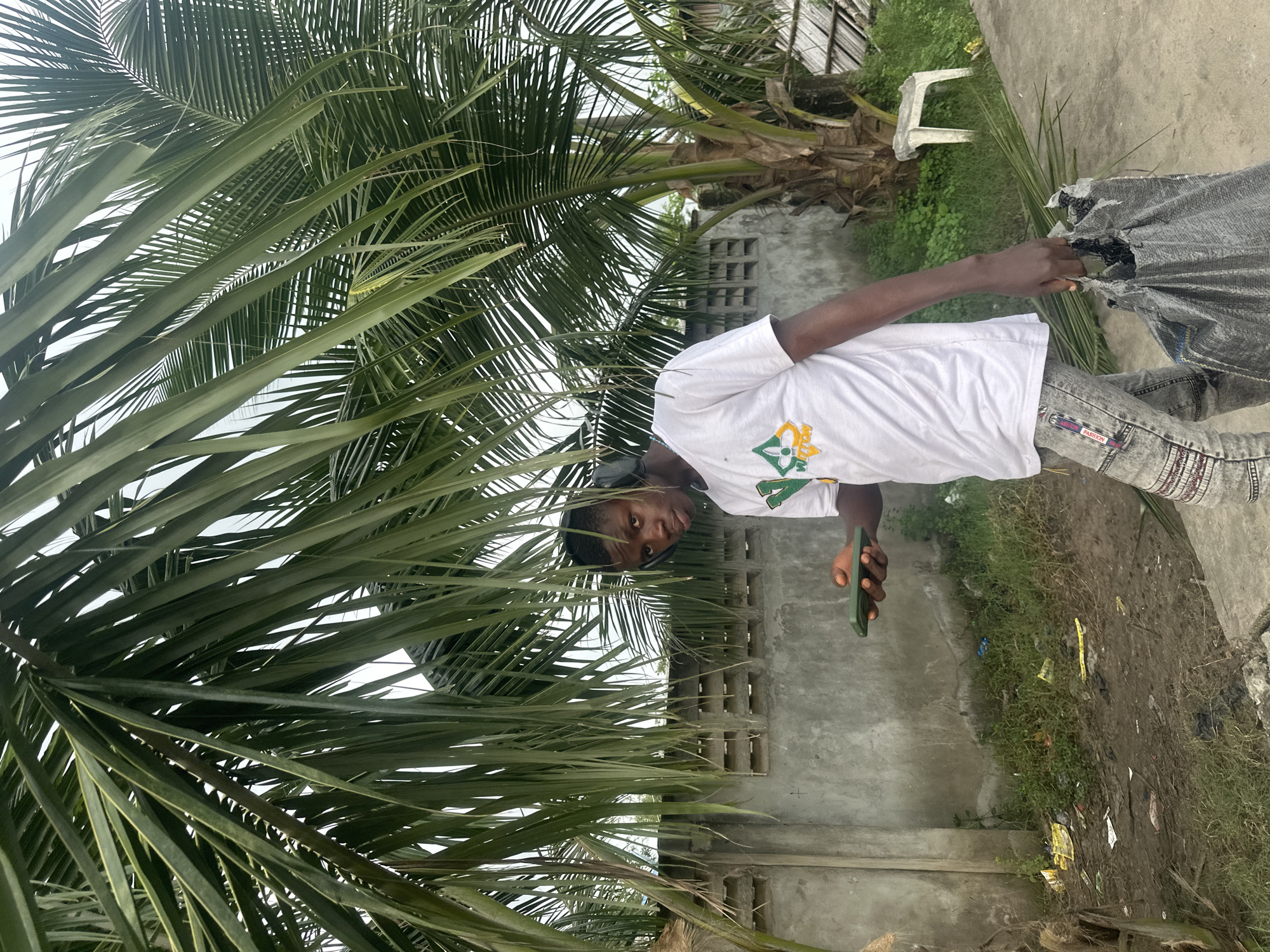 A young Black male is pictured in an outdoor setting, likely in Badagry, Nigeria, given the presence of palm trees and a tropical environment. He is walking towards the viewer's left, holding a green smartphone in his right hand and a torn, dark gray plastic bag in his left. He is wearing a white t-shirt with a green and yellow logo and distressed grey jeans. Black headphones are visible around his neck and ears. The background consists of a rough, grey concrete wall with horizontal ventilation openings, overgrown green grass, and scattered debris on the ground. A palm tree trunk and fronds dominate the upper left and right portions of the image, creating a shaded canopy. A small, white plastic stool is visible in the background near the base of a palm tree. The overall lighting suggests an overcast day.