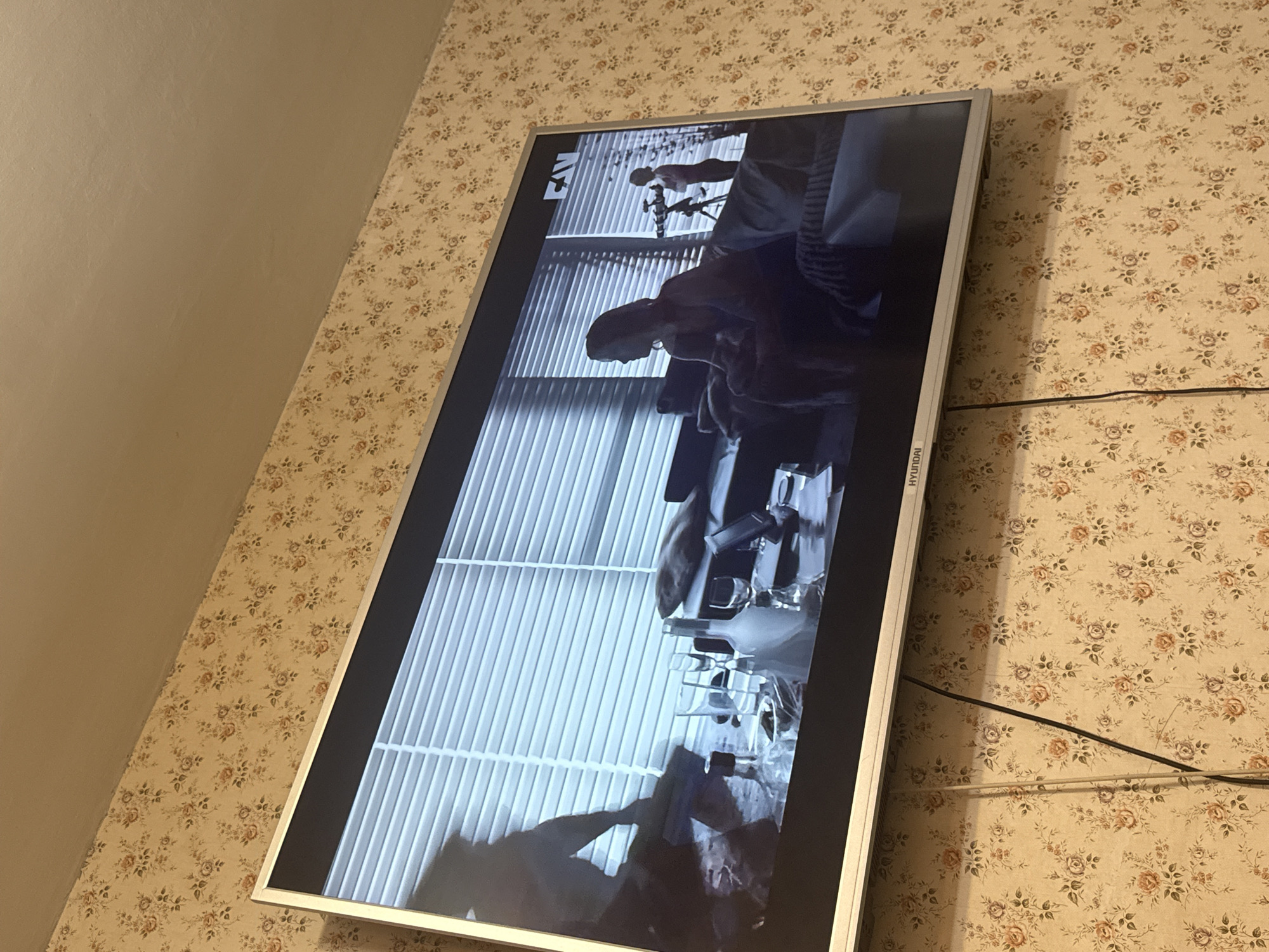 A rectangular television set, bearing the brand name "Hyundai" is mounted on a wall covered in floral wallpaper. The screen displays a black and white image of a person and what appears to be some sort of rack, possibly drying rack. A power cord or other cable runs along the wall adjacent to the television. The lighting suggests an interior setting. There is no information in the image to verify the location in the city of Mytishchi, Russia.