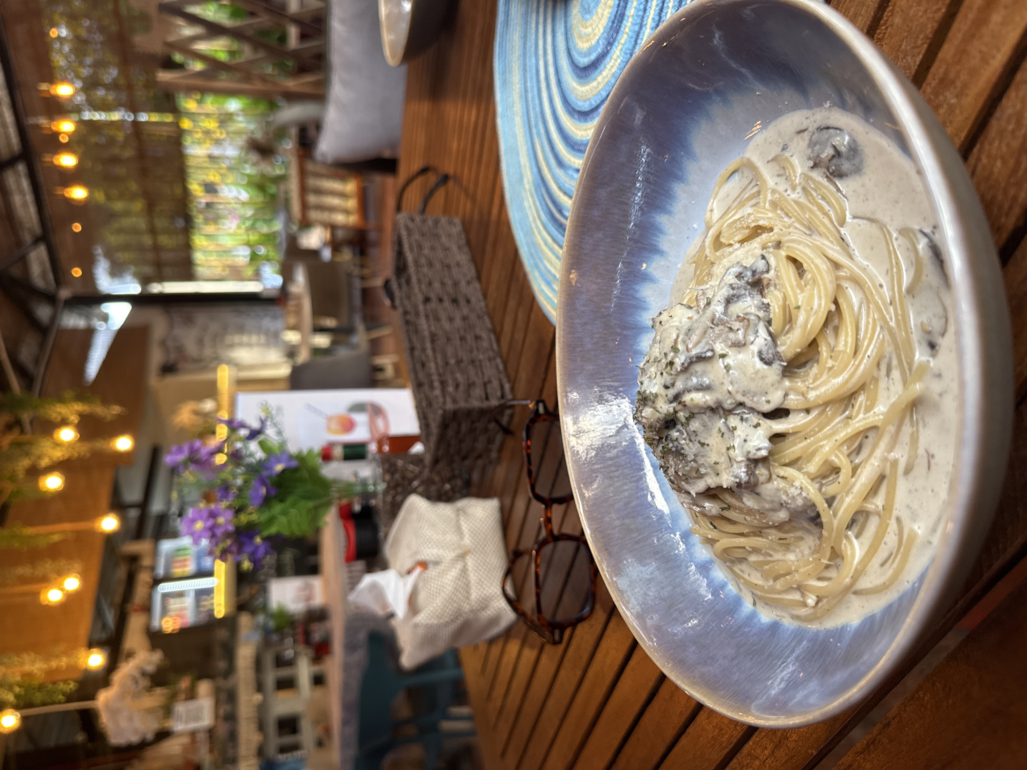 The image shows a plate of spaghetti with a creamy mushroom sauce, possibly truffle, on a wooden table, in what appears to be an outdoor dining area of a restaurant or café. The location may be in Baan Mae Nam, Thailand, based on prior context. The dish is in a blue and white bowl, placed on a round blue and white patterned placemat. Adjacent to the plate is a small brown bag, a pair of tortoiseshell-framed glasses, and a glimpse of flowers. The background is blurred, showing lights, plants, and suggesting an open-air, possibly tourist-oriented, environment. No people are clearly visible.