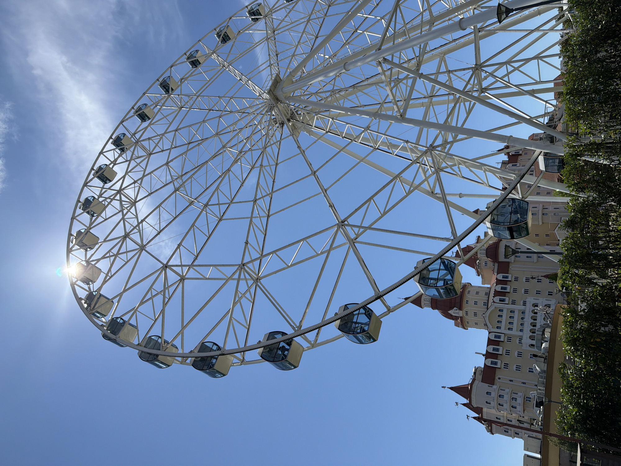 An extremely large, white Ferris wheel dominates the right side of the frame, its intricate metal structure creating a geometric pattern against a bright, clear blue sky. The sun glints brightly from behind the top of the wheel, causing lens flare. Several enclosed gondolas, with tinted blue windows, are attached to the outer rim of the Ferris wheel, some appearing empty. In the lower left and center of the frame, a large, ornate building with red-tiled, conical roofs and numerous windows is visible. The building's facade is a light yellow color with white trim. In the foreground, dark green, manicured bushes partially obscure the view of the building and the base of the Ferris wheel. The overall impression is a sunny day at an amusement park or resort. The location is identified as the city of Sirius, Russia.