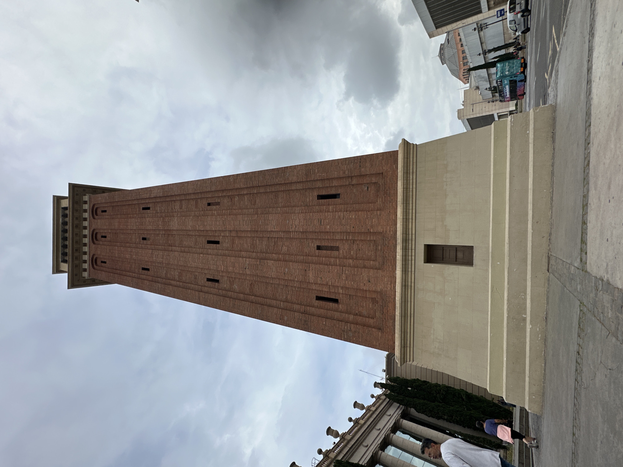 The image shows the Venetian Towers on Avinguda de la Reina Maria Cristina in Barcelona, Spain. The red brick tower is the closer of the two towers. The building that is located in the bottom of the image has a white top with tall glass windows and black columns. There are a few people walking on the sidewalk in front of the building. The sky is overcast with gray clouds.