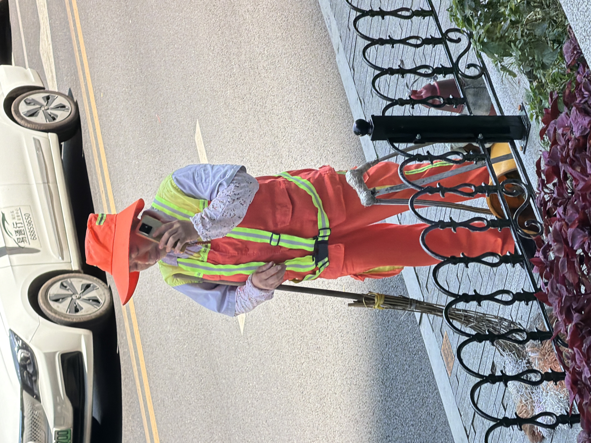 In the Yuzhong District of China, a street cleaner wearing an orange hat and vest with reflective stripes is standing on the sidewalk, speaking on a mobile phone. They are also holding a broom. Behind the cleaner, a white car with a license plate starting with "99" is parked on the street. The environment appears to be an urban setting with a paved sidewalk and road, separated by a decorative black metal fence. The weather is sunny.