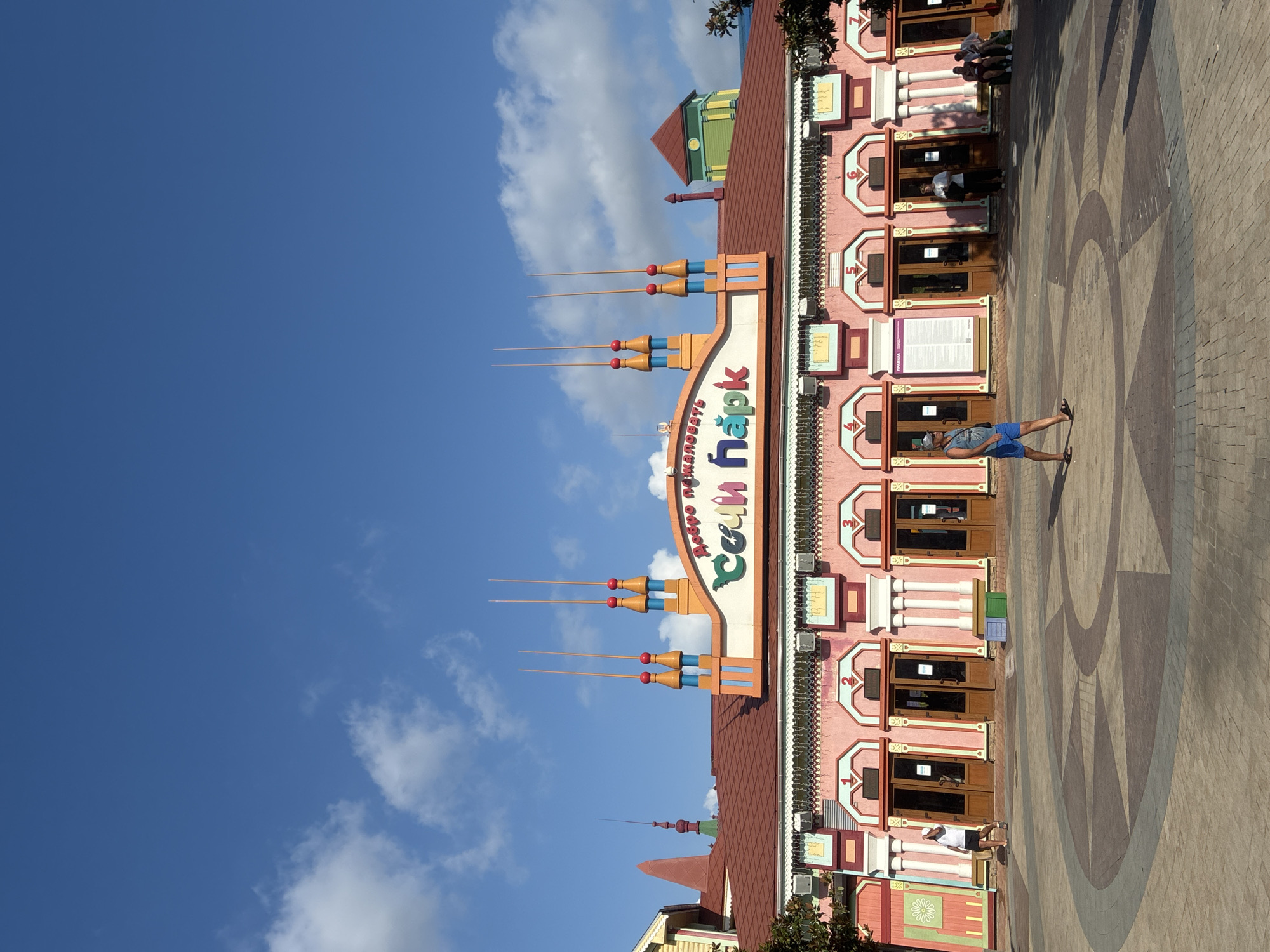 The image shows the entrance to "Sochi Park" in Sochi, Russia. The building is painted pink with colorful decorative elements, including spires on the roof. The sign above the entrance reads "Добро пожаловать в Сочи Парк," which translates to "Welcome to Sochi Park." Several people are visible in front of the building, some walking and others sitting on benches. The ground in front of the entrance is paved with a circular geometric pattern. The sky is blue with some scattered clouds.