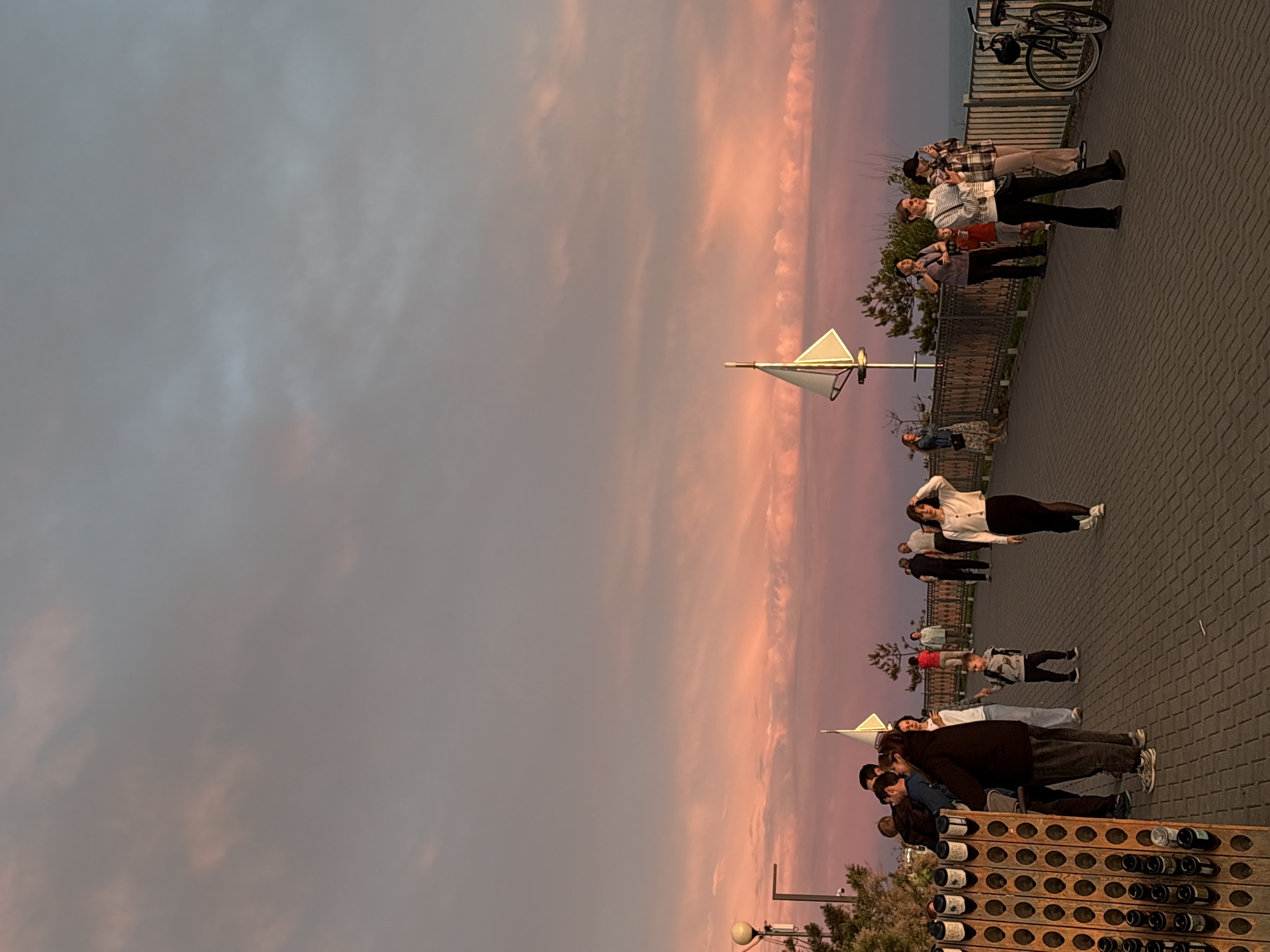 The image captures an outdoor promenade in Starotitarovskaya, Russia, during sunset or twilight hours, characterized by a strikingly colorful sky. The upper portion of the sky transitions from a soft grey-blue to vibrant pink and orange hues, with scattered clouds reflecting the fading light.
Numerous people are present on the paved walkway, which features a herringbone pattern. In the mid-ground, a woman in a white cardigan and dark pants stands facing forward with her hands behind her head. To her left, near a metal railing, a group of individuals is gathered, including a woman in a plaid shirt and another person wearing light-colored pants. A bicycle with a helmet attached is propped against the railing further back.
On the foreground to the left, a large, dark wooden structure with multiple circular cutouts, possibly serving as a decorative screen or a rack for bottles, is prominently displayed. A group of people, including a man in a dark jacket and a child, are standing near this structure. Other figures are scattered throughout the scene, some appearing to stroll or stand idly.
Distinctive white, triangular sail-like structures, possibly modern streetlights or art installations, are visible along the walkway. Trees and bushes line the railing, which separates the promenade from a body of water visible in the background. The overall mood is serene and relaxed, suggesting people are enjoying an evening walk. No discernible text is visible in the image.