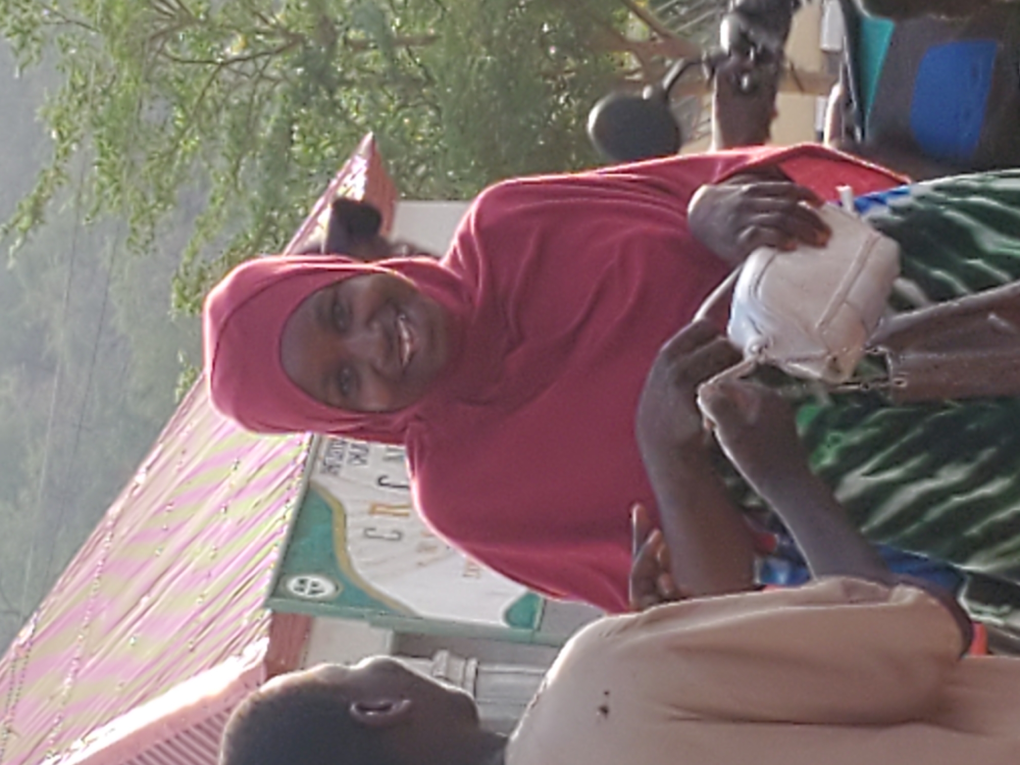 This outdoor photograph, taken in Mararaba, Nigeria, captures a lively daytime scene featuring multiple individuals engaged in an interaction. The central figure is a woman with dark skin, positioned in the middle-right of the frame, wearing a bright red hijab and a matching top. She is smiling warmly and looking directly towards the camera, conveying a friendly and approachable demeanor.
Her hands are holding a small, white, somewhat squarish object, resembling a plastic container or a compact bag. Another person's dark-skinned arm, adorned with green striped fabric, reaches towards this white object from the lower right, suggesting an exchange or shared interaction over the item. In the foreground, partially visible, is the head and shoulders of a third individual, wearing a light-colored garment, facing the smiling woman. Further in the background, another person is seen near what appears to be a motorcycle or scooter.
The setting is outdoors, characterized by abundant green foliage and trees in the upper background. To the left, a distinctive roof structure with diagonal pink and light green stripes adds a vibrant element to the scene. Behind the central woman, there's a light-colored surface, possibly a wall or sign, with some faint, largely illegible text, vaguely showing letter-like forms such as 'C R C J'. The clear, natural lighting indicates it is daytime. The overall impression is one of positive social interaction and community engagement.