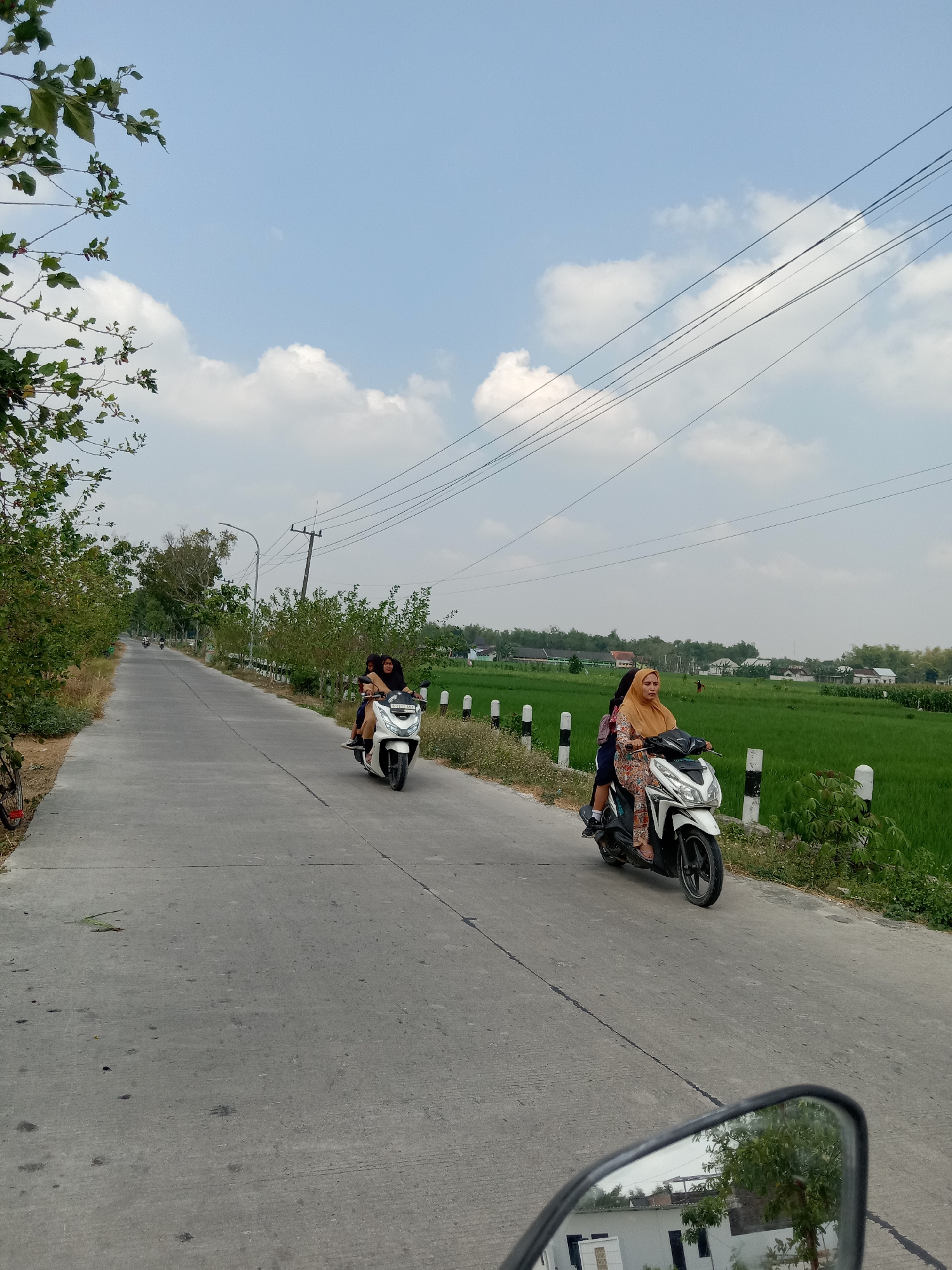 The image depicts a rural road in Sidobandung, Indonesia, under a bright, partly cloudy sky. Two motorcycles are traveling on the paved road. The first motorcycle, closer to the foreground, carries two individuals. The rider is wearing a light-colored, patterned outfit and a yellow headscarf. The passenger behind is wearing a dark hijab. The second motorcycle, further down the road and slightly to the left, also has two occupants, both wearing dark clothing and hijabs.
The road is bordered by lush green rice fields on the right and vegetation, including trees and bushes, on the left. Several utility poles and power lines are visible overhead. In the distance, buildings can be seen interspersed with more greenery. The overall impression is of a sunny day in a rural Indonesian setting. The perspective suggests the photo was taken from another moving vehicle on the same road.