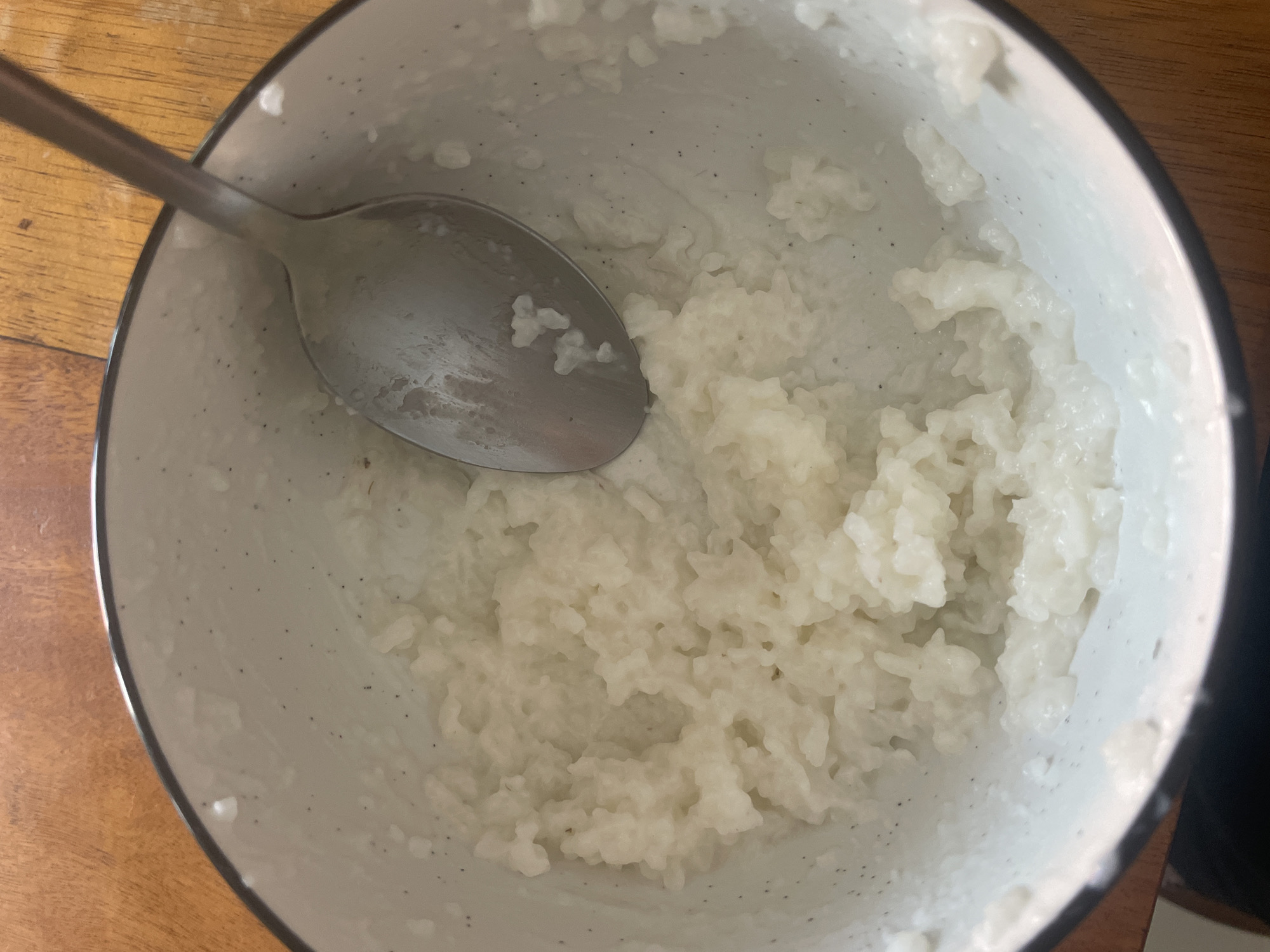 A top-down view of a speckled white bowl containing a portion of cooked rice, with a metal spoon resting inside. The bowl sits on a wooden surface. The photograph was likely taken indoors. The exact location in Denpasar, Indonesia cannot be determined from the image alone.