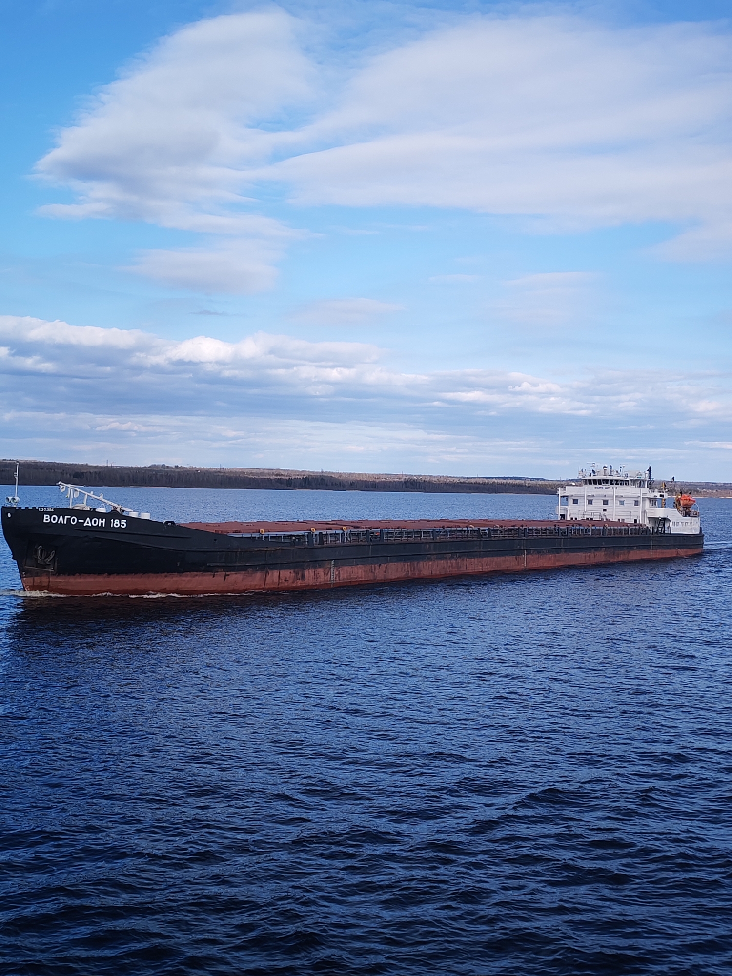 The image shows a large cargo ship, identified as "ВОЛГО-ДОН 185" (Volgo-Don 185), navigating a wide body of water under a partially cloudy blue sky. The vessel is a dark-hulled bulk carrier with a reddish-brown waterline, appearing to be laden with cargo, possibly gravel or another bulk material, visible in its open holds. Its superstructure, located at the stern, is white and features multiple levels, windows, and navigation equipment, including an orange life raft mounted on the side. The ship is moving from left to right, creating a distinct wake behind it and at its bow.
The water is a deep blue, showing ripples and small waves. In the distance, a low, forested shoreline stretches across the horizon, suggesting a large river or canal. The sky above is clear blue in many parts, with scattered white and light gray cumulus clouds. The overall lighting indicates it is daytime, likely late morning or early afternoon, with clear visibility.
The most prominent text is "ВОЛГО-ДОН 185" painted in large white Cyrillic letters on the ship's black bow. A smaller number, "130364," is also visible near the bow above the main text. The name "Volgo-Don" is indicative of its operation in the Volga-Don region of Russia, consistent with the provided location context of Novaya Svobodka, Russia. The scene portrays routine commercial navigation on an inland waterway. No individuals are clearly visible on the ship or elsewhere in the frame.