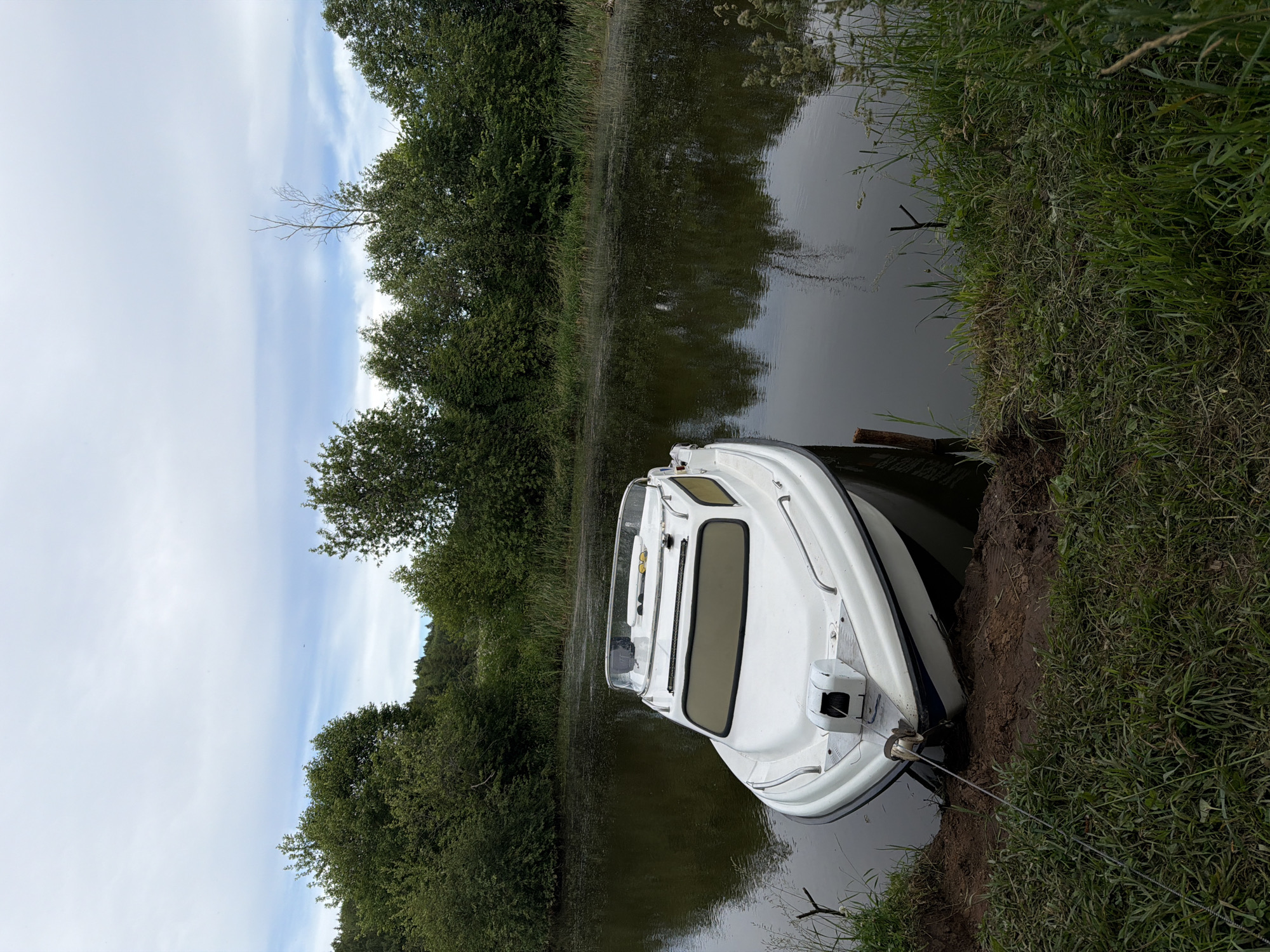 A white boat is partially grounded on a muddy bank beside a calm body of water. The boat is oriented towards the right of the frame, with its bow facing slightly left. Green foliage, including trees and grass, lines the banks of the water, creating a natural landscape. The sky above is overcast with scattered clouds. The scene appears to be in a rural or natural setting, possibly on a river or lake. The location is identified as Garevaya, Russia.