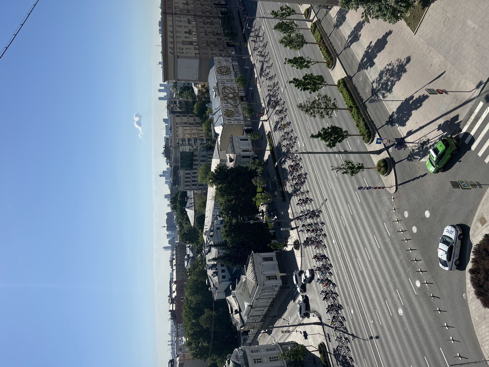 A high-angle, daytime shot depicts a city street lined with trees and buildings. The sky is a clear blue with a wisp of cloud in the upper left. A line of cyclists extends along the street, passing various buildings. A green and white car is parked on the right side of the street at a crosswalk. A white police car is parked further down the street. Shadows of the trees fall across the street. The city is Unknown, Unknown.