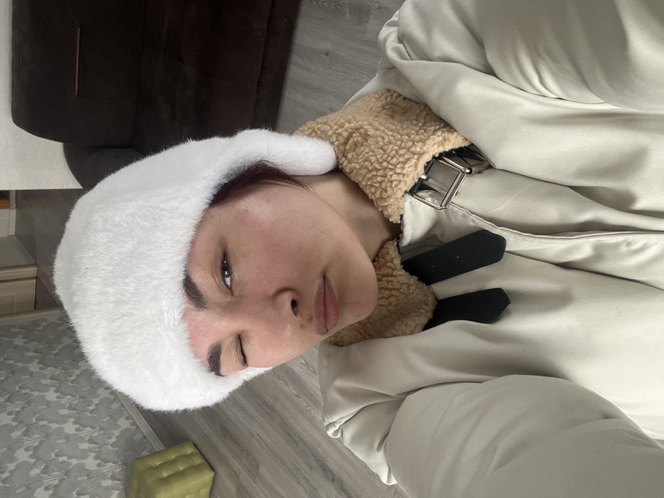 This image depicts a single individual within an indoor residential setting in Starobaltachevo, Russia. The individual, appearing to be an adult female, is centered and occupies the majority of the frame, captured in a selfie-style, upward-angled perspective. She has dark hair, dark eyebrows, and her left eye is closed in a distinct wink, while her lips are pursed forward. She is wearing a white, fluffy winter hat, covering her head and ears. Her attire also includes a light-colored, possibly off-white or beige, puffer jacket with a prominent light brown, sherpa-style collar. A metallic silver-colored buckle and two black strap elements are visible on the right side of the jacket's chest area. The background elements include a light wood-toned floor. To the left, a wall is covered with a light gray, abstract, cloud-like patterned wallpaper, with a light green upholstered cube or ottoman resting on the floor against it. To the right, a section of a dark brown upholstered sofa is visible. A doorway with a light-colored frame is partially visible in the upper left background. No other individuals or interactions are depicted.