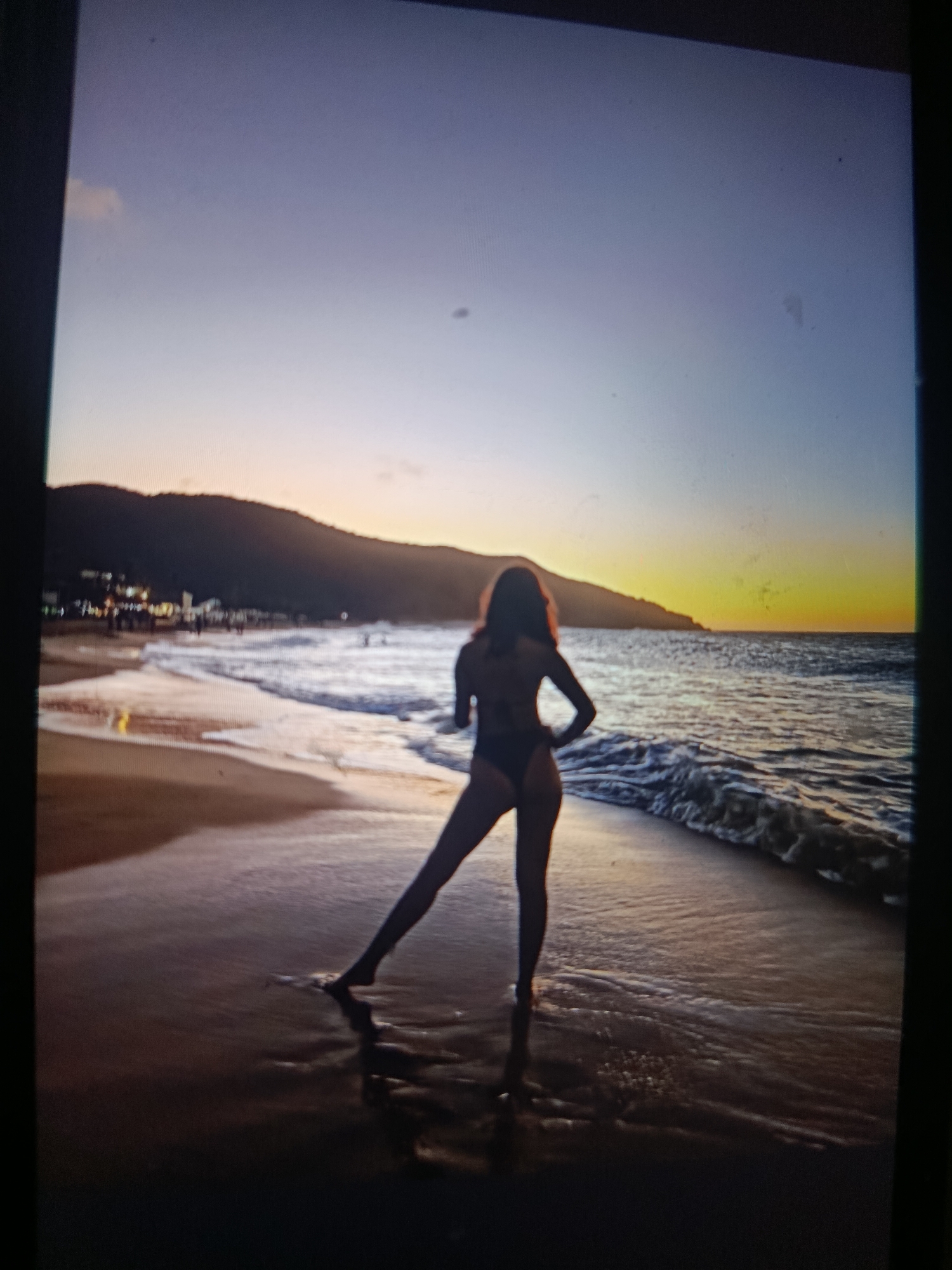 The image captures a silhouette of a woman standing on a beach at sunset in Juan Griego, Venezuela. The woman is wearing a dark swimsuit, with one leg extended out to the side. She is facing the ocean, with her hands on her hips. The sun is setting over the water, casting a warm, golden light on the scene. Waves are gently rolling onto the beach. In the background, there is a silhouette of a mountain and some lights from the town of Juan Griego.