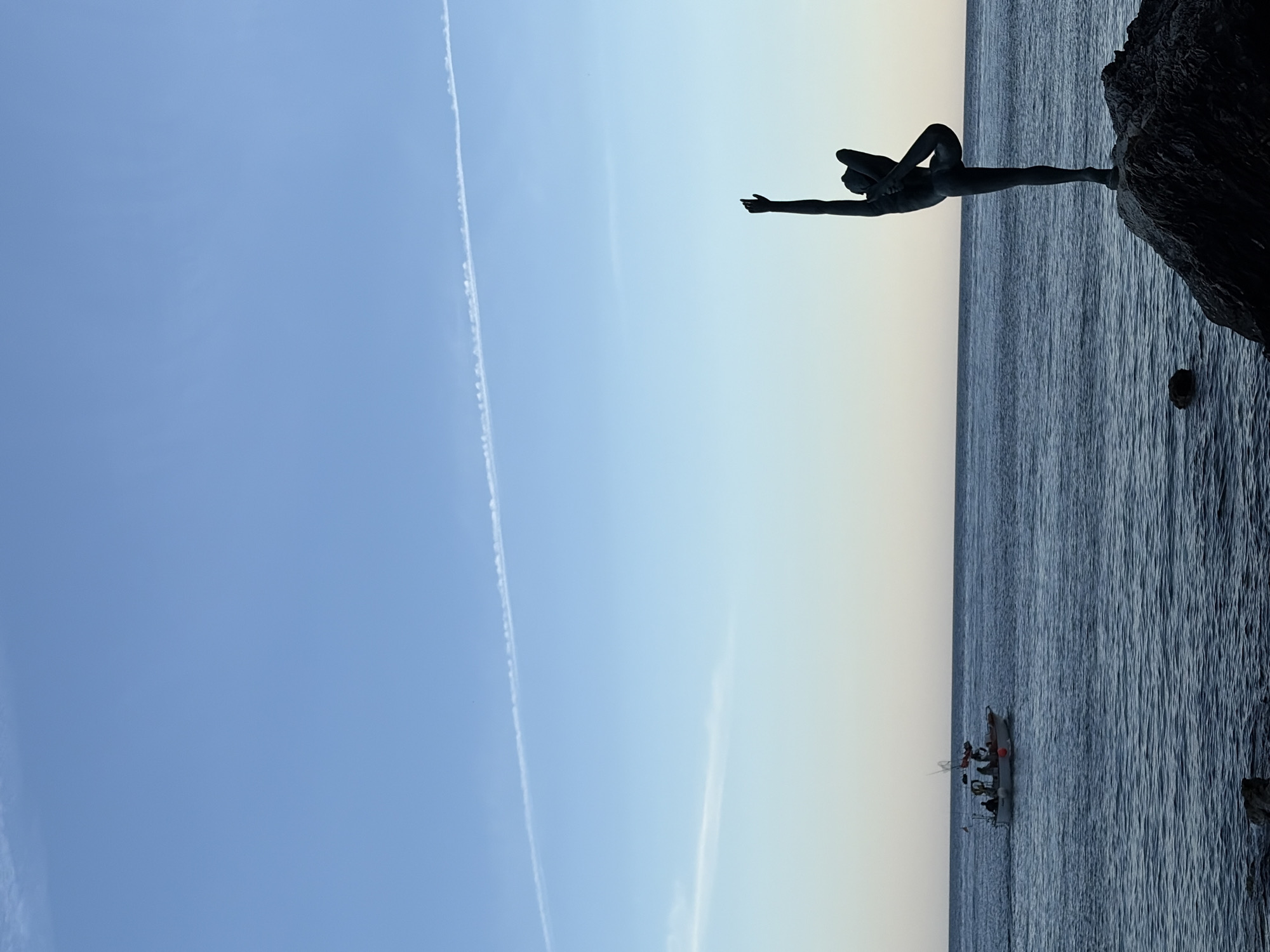 The landscape image depicts a serene coastal scene, likely captured during dawn or dusk, dominated by the silhouette of a graceful statue overlooking the sea. The statue, a slender human figure, is captured in an elegant, almost acrobatic pose, balancing on one leg with the other bent upwards and held by one hand, while the opposite arm extends forward. It stands prominently atop a dark, rugged rock formation located on the right side of the frame. This figure strongly resembles the "Dancer of Budva" (Budvanska balerina) statue, located in Budva, Montenegro.
The sky above the horizon transitions from a light, warm yellowish-blue near the sea to a clearer, deeper blue higher up, indicating the soft light of either early morning or late afternoon. Two faint, thin contrails are visible cutting across the vast, clear expanse of the sky.
The sea occupies the lower and middle portions of the image, presenting a gently rippled surface under the soft, ambient light. Further out on the water, towards the left side of the frame, a small boat is visible with several people onboard, suggesting a leisurely activity such as fishing or sightseeing. The overall weather appears calm and clear. No text is visible in the image.