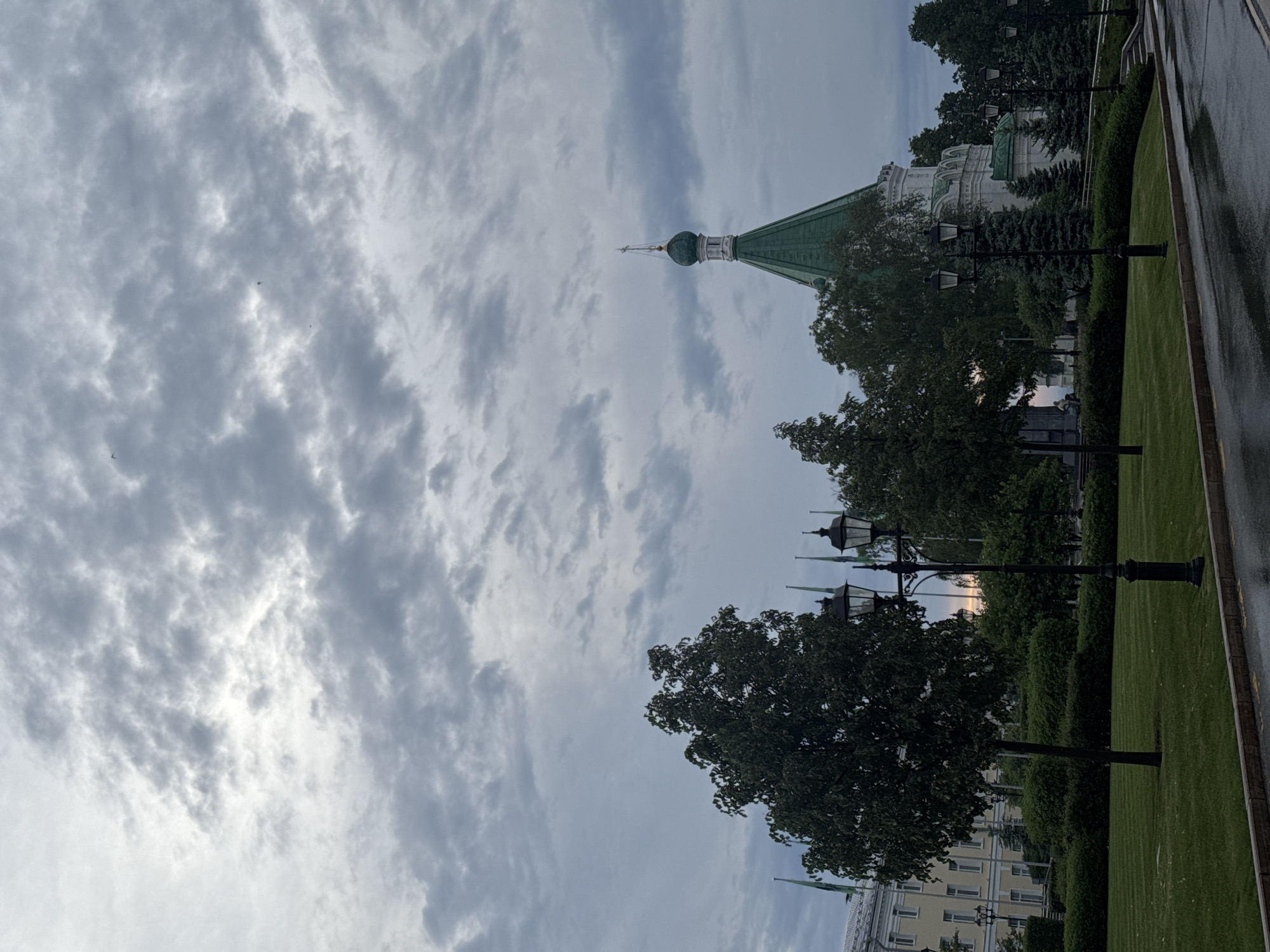 The image shows the Alexander Nevsky Cathedral in Nizhny Novgorod, Russia, taken from ground level. The cathedral's green roof and dome are visible, surrounded by trees and lampposts. A building appears to be behind the trees, with a cloudy sky above. Wet pavement is visible in the lower right.