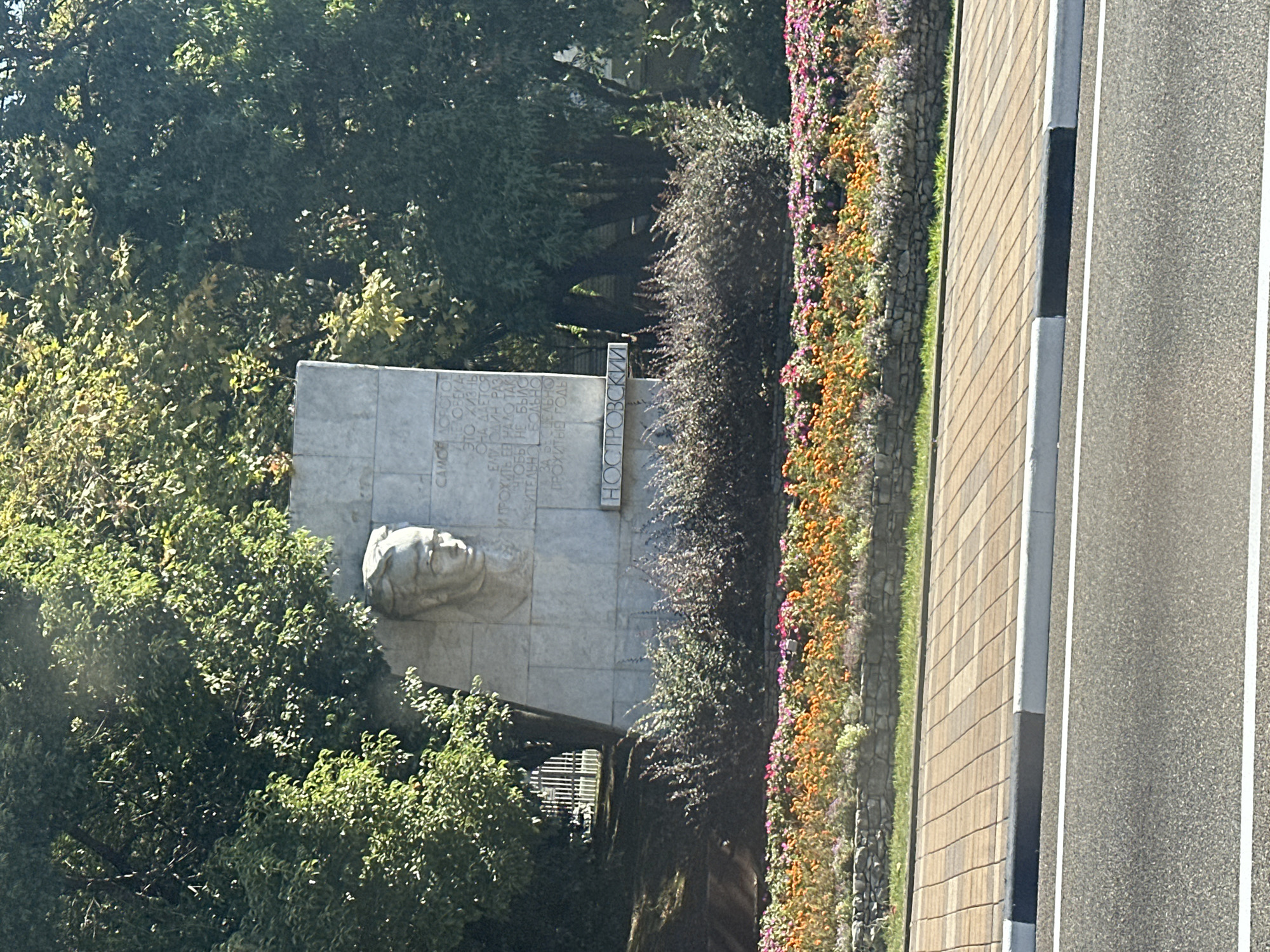 This is a medium-angle, eye-level, full shot of a monument with a bas-relief of a man's head on a light-colored stone slab. The monument is located in front of a dense cluster of green trees, some of which are casting shadows. Below the monument, a dense bed of dark green shrubs is visible, which in turn is in front of a vibrant flower bed with orange, pink, and purple flowers. The flower bed is bordered by a low stone wall. In the foreground, a paved sidewalk made of rectangular stones leads to a road with white lane markings. The entire scene appears to be outdoors in an urban setting, likely a park or public square. The inscription on the monument appears to be in Russian. This monument is likely located in the city of Sochi, Russia, as indicated by the inscription, which reads "ОСТРОВСКИЙ," a reference to Alexander Ostrovsky, a famous Russian playwright.