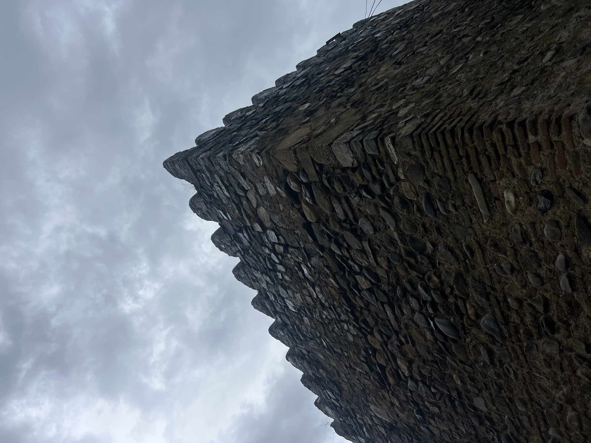 The image shows a low-angle shot of a stone tower against a cloudy sky. The tower is constructed with irregular stones and appears to be old. The sky is overcast, with a mix of gray clouds. The perspective emphasizes the height and texture of the tower. There are no people or other discernible objects in the frame, apart from a few sparse blades of grass growing from the top of the tower and some wires on the right side. The image is a straightforward depiction of an architectural element in an outdoor setting. The location of Shalikiantkari, Georgia, is mentioned in the prompt but not visually identifiable from the image itself.