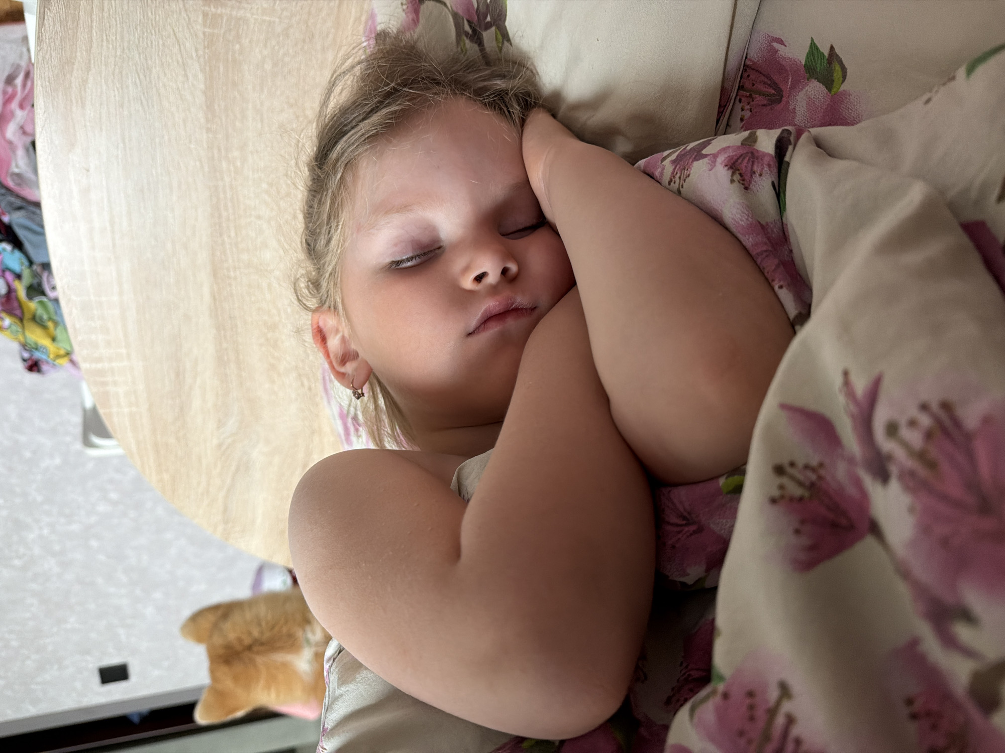 This close-up image captures a young child, likely a girl, sleeping peacefully indoors in Chaikovsky, Russia. Her face is serene, with eyes closed and lips slightly parted in slumber. She has light-colored hair and wears a small star-shaped earring in her left ear. Her arms are bent and tucked close to her body, with one hand resting near her forehead.
She is partly covered by bedding featuring a distinct floral pattern of pink and purple flowers with green leaves. To the left of her head, a light-colored wooden surface, possibly a table, is visible. In the bottom-left background, slightly out of focus, a small, golden-brown object, possibly a pet like a cat or dog, or a soft toy, can be seen.
The soft, natural lighting suggests a quiet moment of rest, perhaps a daytime nap. The overall atmosphere is tranquil and calm, emphasizing the child's peaceful sleep. There is no visible text in the image.