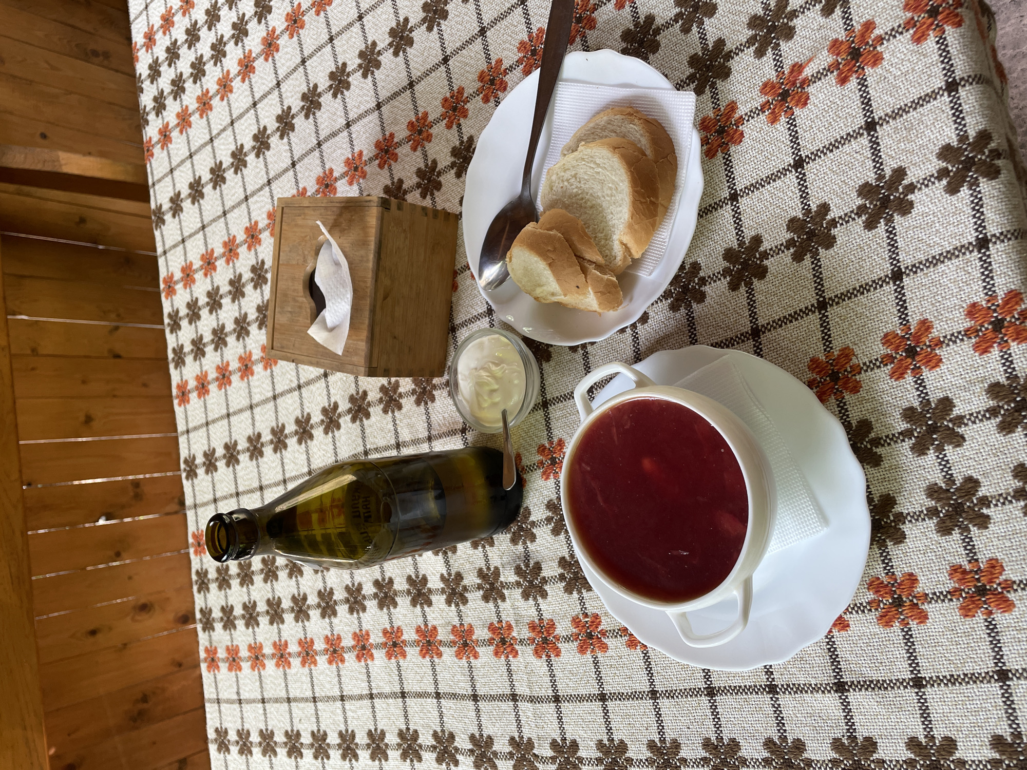 A high-angle shot captures a rustic tabletop meal set against a wooden background, likely in Kamianytsia, Ukraine. The central focus is a white bowl filled with a rich, dark red soup, characteristic of borscht, resting on a matching saucer. Beside it, a small glass contains a dollop of white cream, presumably sour cream or smetana, with a small spoon.
To the left, a dark green bottle lies horizontally on the tablecloth. Partially visible Cyrillic text on the bottle clearly shows the word "НАПІЙ," meaning "beverage" or "drink." Further up, a white plate holds several slices of white bread, accompanied by a silver spoon. A wooden tissue box with a single white tissue protruding from it is positioned near the bread.
The table is covered by a textured beige and white tablecloth featuring a distinctive pattern of brown and orange floral motifs arranged in a grid-like fashion, reminiscent of traditional folk design. The edges of the table and the background reveal warm-toned wooden planks, suggesting a cozy, possibly outdoor or cabin-like setting. The natural lighting indicates it is daytime. The overall impression is a simple, traditional meal, perhaps lunch or dinner, served in a relaxed environment consistent with the Ukrainian context.