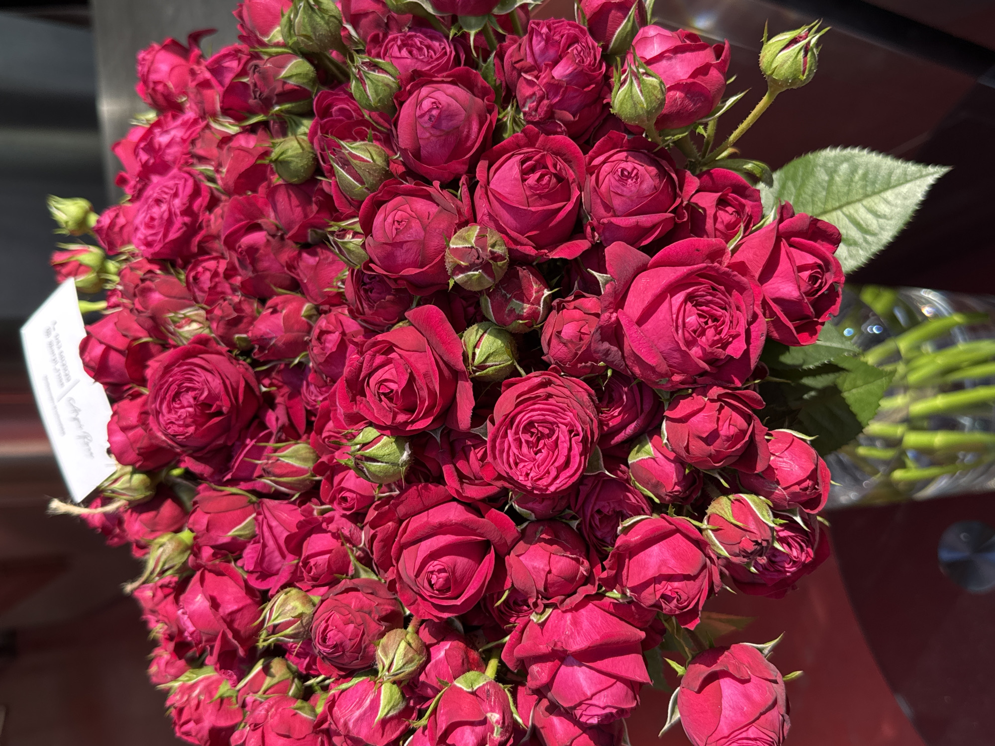 A bouquet of red roses is densely packed together. Some roses are fully bloomed, while others are still buds. Green leaves are visible amongst the roses. In the background, the stems of the roses are seen in a glass vase. A white tag is partially visible on the left side of the bouquet. The setting is Yerevan, Armenia.