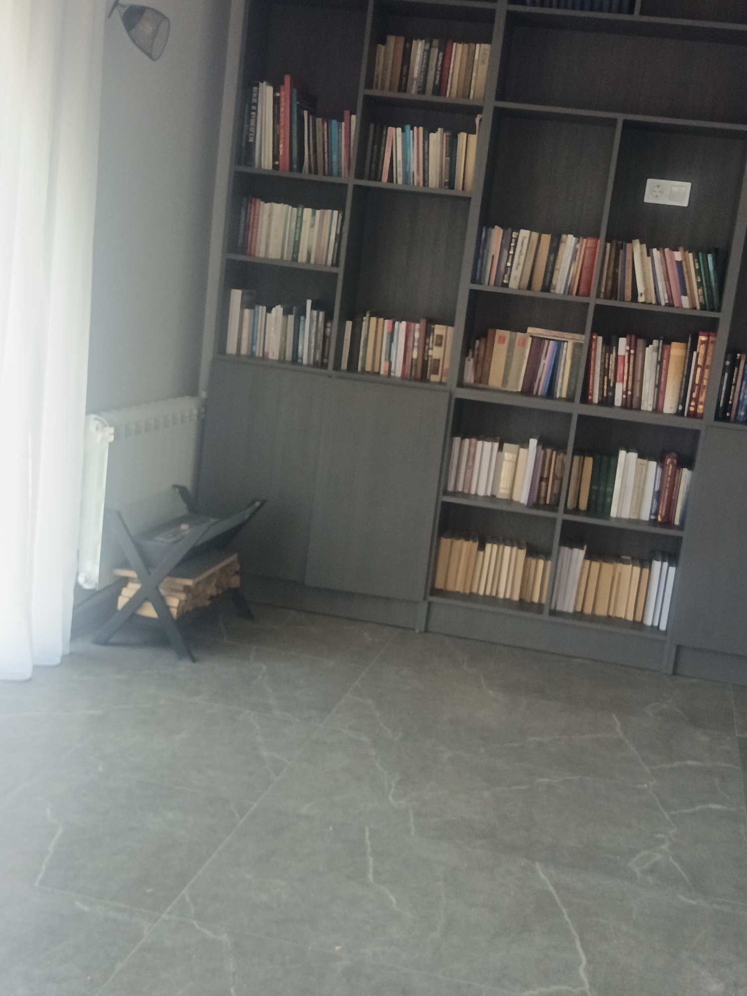 This is a medium-shot, eye-level, indoor, full-color photograph taken in what appears to be a living room or study. The scene is dominated by a large, dark gray, built-in bookshelf that takes up a significant portion of the right side of the frame. The bookshelf is filled with numerous books of varying sizes and colors, neatly arranged on multiple shelves. A white electrical outlet is visible on the wall behind the books on one of the shelves.
To the left of the bookshelf, a white sheer curtain hangs, covering a window or wall. In front of the curtain and to the left of the bookshelf, there is a white radiator. Below the radiator and to the left, a black, modern-looking log holder with firewood stacked inside is positioned on the floor. The floor itself is a gray tiled surface with a subtle marble-like pattern. A light fixture with a metal arm is attached to the wall near the top left corner of the image, emitting a soft light. The overall ambiance is one of quiet sophistication and organization.