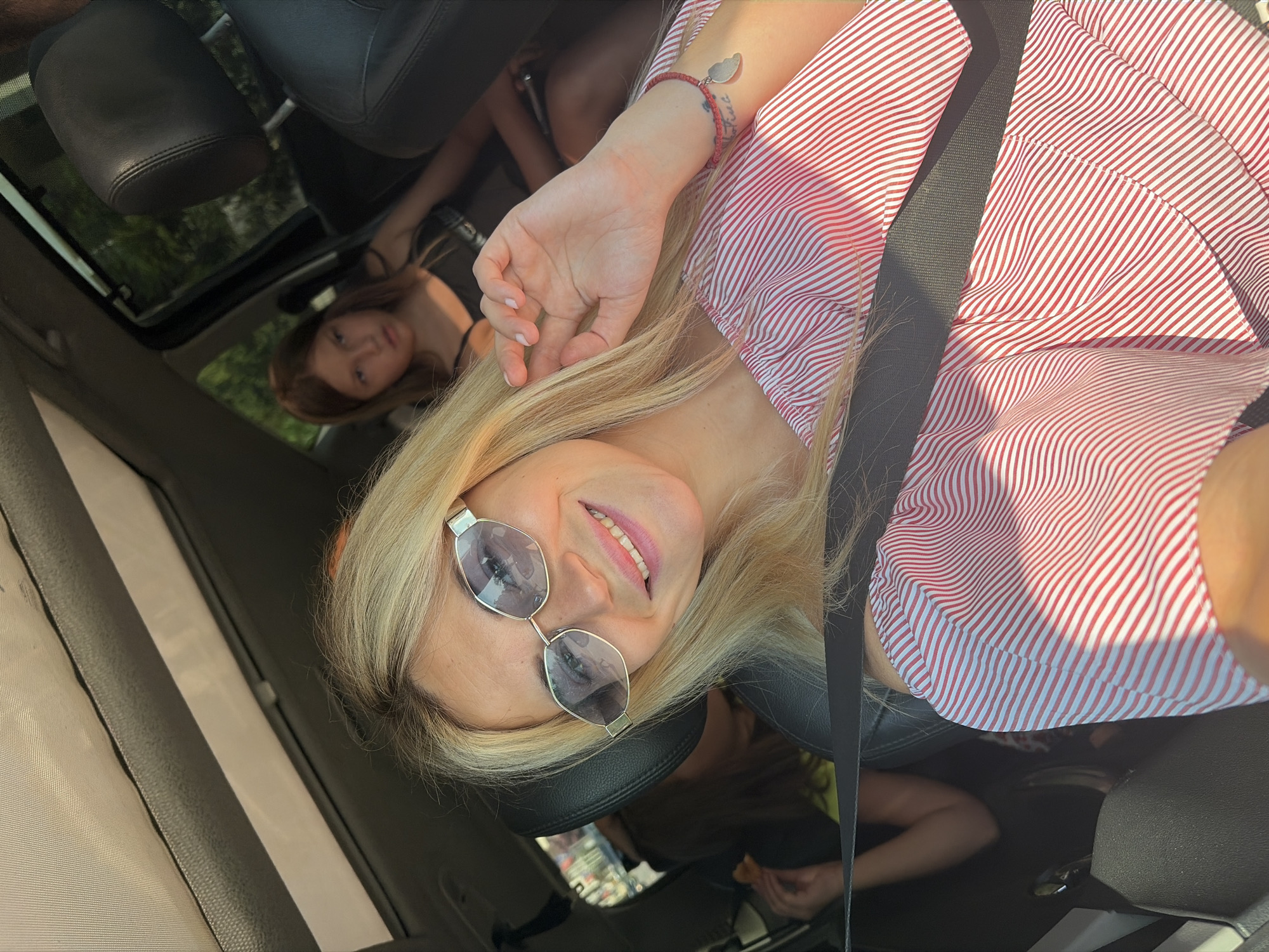 A blonde woman wearing sunglasses and a red and white striped off-the-shoulder top is taking a selfie inside a car. She is smiling and has her right hand raised to her face. Behind her, in the back seat, a young girl with brown hair is visible, looking away from the camera. Another person, partially obscured, is also in the back seat. The interior of the car is visible, including the headrests and seatbelts. The scene appears to be a sunny day, with light filtering into the car. The location is the city of Budva, Montenegro.