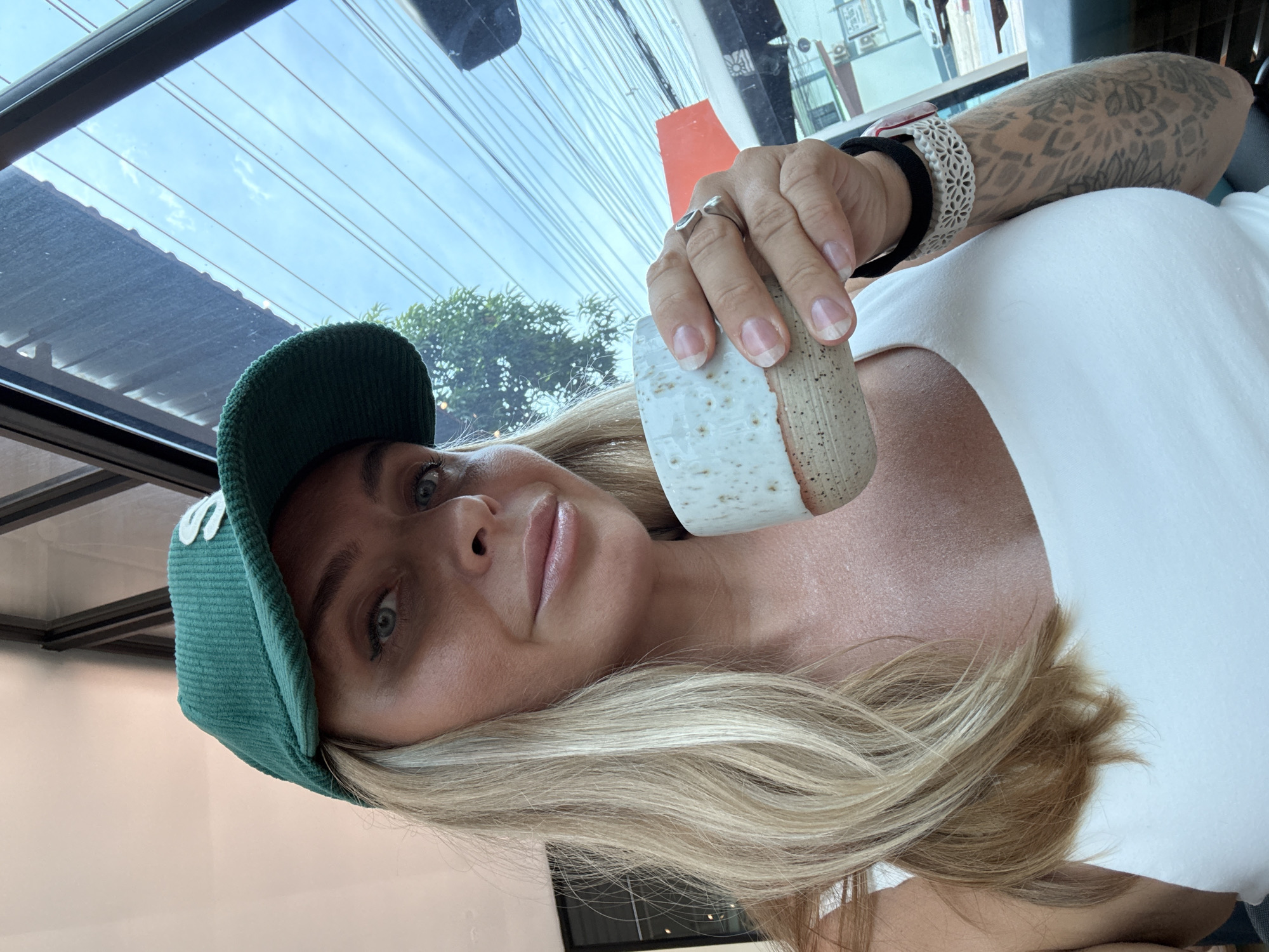 In Ko Samui District, Thailand, a woman with blonde hair and blue eyes, wearing a green corduroy baseball cap and a white tank top, holds a speckled ceramic mug of coffee. She has a ring on her right hand and a tattoo on her left forearm. The background shows a window with a view of tangled electrical wires, trees, and a bus.