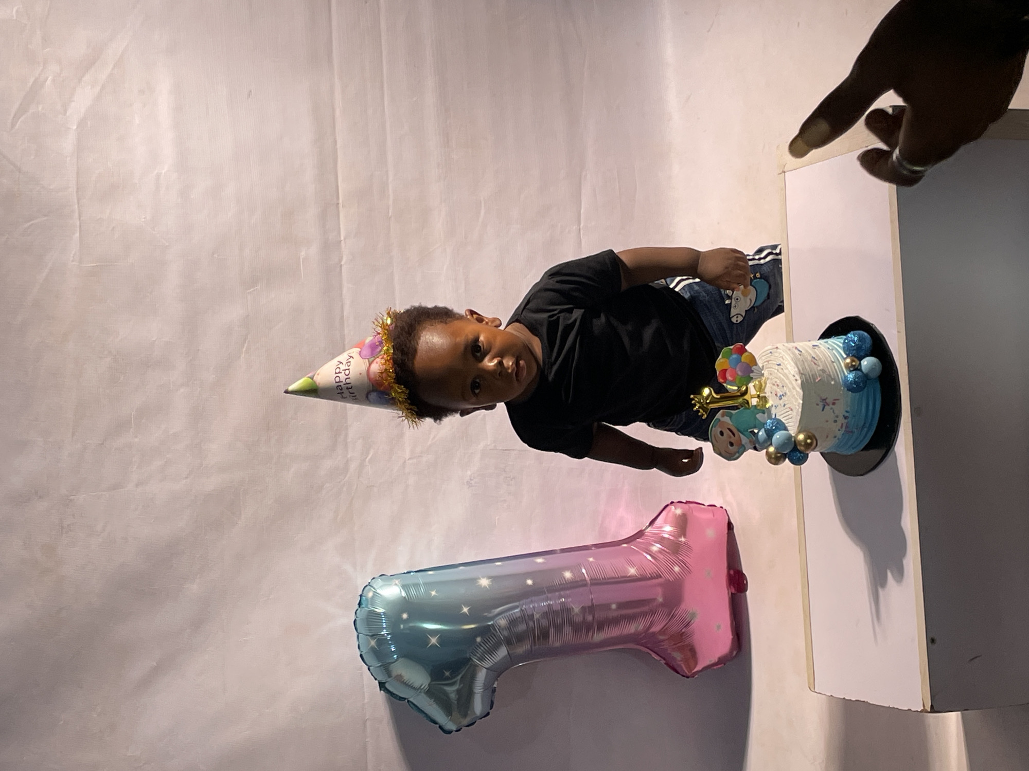 A child wearing a birthday hat is lying on the ground next to a number one shaped balloon. A birthday cake sits on a pedestal next to the child. A hand reaches in from the upper right side of the image towards the cake. The setting is an indoor studio with a white backdrop. The exact location in the city of Barakin Akawo, Nigeria is not discernible from the image.