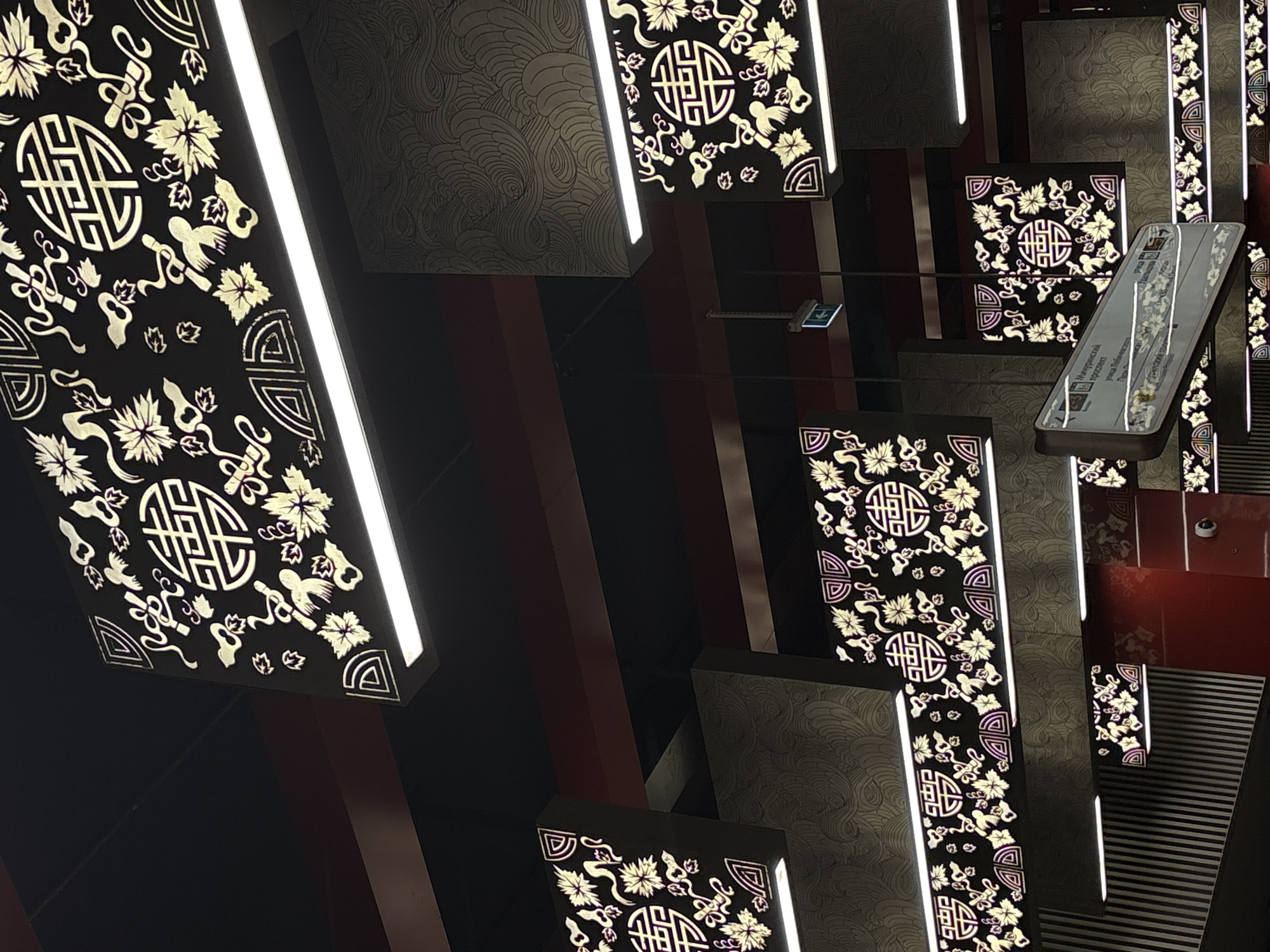 The image shows a section of the interior of the Arbatskaya Metro Station in Moscow, Russia. The view is tilted upwards, showcasing the station's ornate ceiling design. The ceiling is adorned with a series of rectangular panels. Some of these panels feature traditional Asian-inspired patterns of flowers and geometric symbols against a black background, accentuated by integrated linear lights. Other panels display a swirled, textured pattern. There is also a sign with text written in Russian. The overall atmosphere is a blend of grandeur and intricate detail.