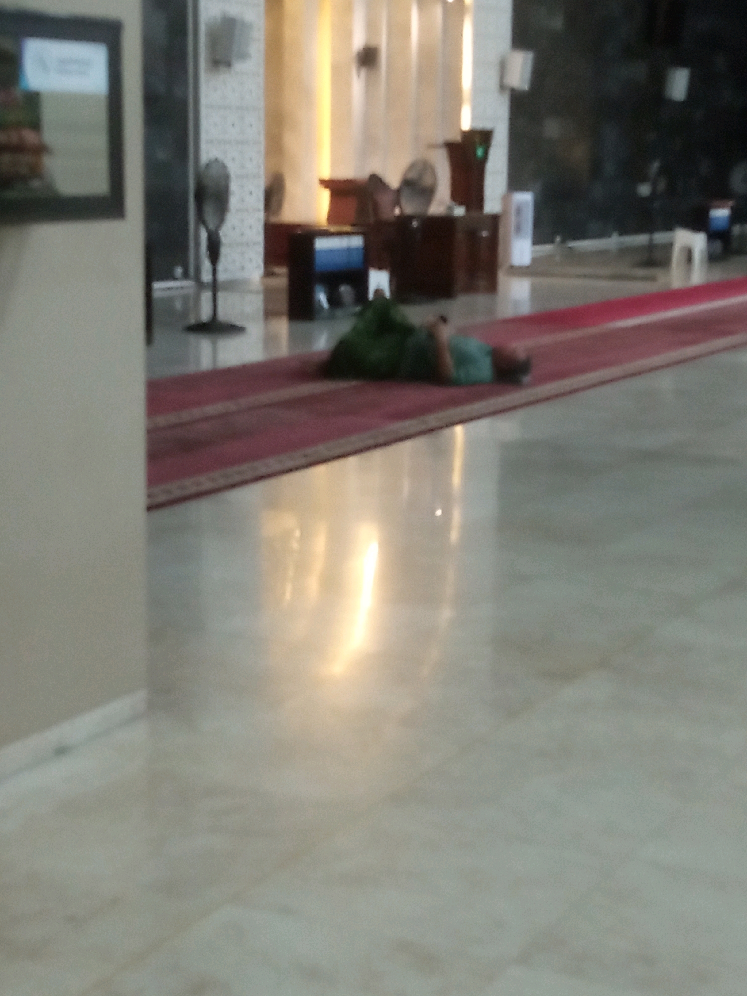 The image depicts an indoor scene, possibly a public building or a place of worship, with a person lying down on a red carpet. The flooring appears to be polished tile, reflecting the ambient light. In the background, there are fans, furniture, and what seem to be architectural elements of the building. The lighting is warm and diffused, suggesting artificial illumination. The person is wearing dark clothing and is positioned on their side, appearing to be resting or sleeping. The overall scene is somewhat blurry, possibly due to motion or low light conditions. Without further information or context, the specific location in Bontang, Indonesia, cannot be determined.