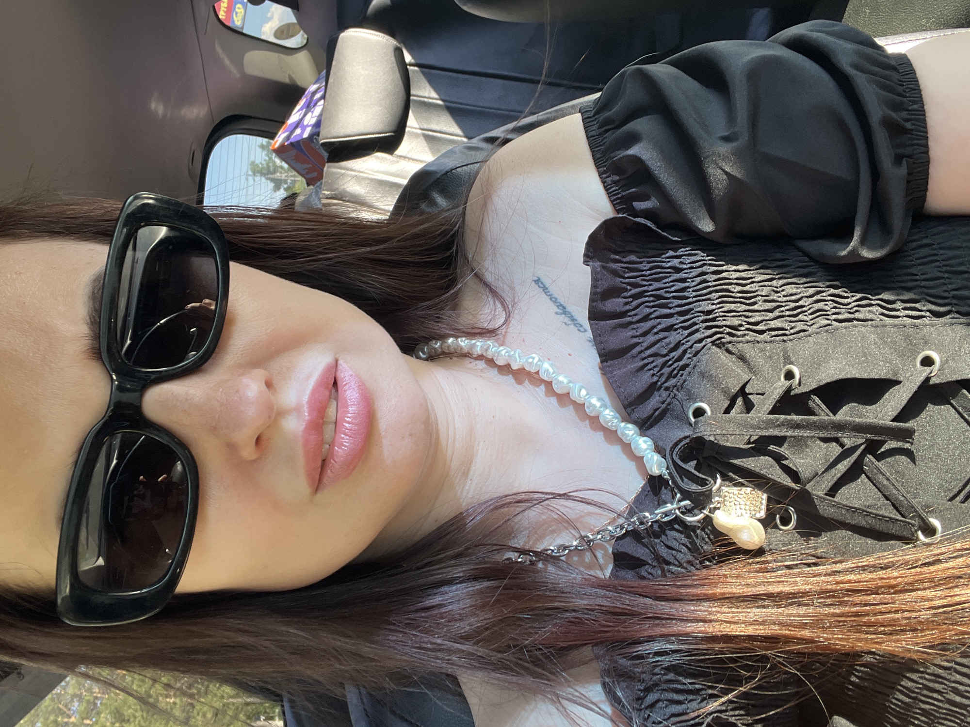 A close-up, high-angle portrait shows a person, likely a woman, reclining or posing inside a vehicle during a sunny day. The subject's long, dark brown hair, highlighted with reddish tones towards the ends, is spread across her chest and shoulder. She wears large, black, rectangular sunglasses that obscure her eyes, and her lips are slightly parted, revealing a glossy, light pink or peachy lipstick.
She is dressed in a black top or dress characterized by a smocked, textured bodice with a lace-up front featuring silver eyelets and a tie. Her sleeves are full and gathered, contributing to the garment's voluminous silhouette. Around her neck, she wears a necklace composed of irregularly shaped pearls. A silver-toned chain extends from the pearl necklace, holding two charms: one appears to be a small, light-colored shell or baroque pearl, and the other is a rectangular, possibly studded, metallic piece. On her upper chest, near the collarbone, a script tattoo is partially visible, with legible fragments that suggest the word "estamos."
The setting is inside a car or similar vehicle, with dark upholstery visible in the background. Bright sunlight streams through the windows, creating reflections of outdoor scenery including trees and a clear sky. A segment of a rearview mirror is also visible. In one reflection, some red text, possibly "IPSAO" or "IPSA," along with numbers like "150" or "190," can be faintly seen, likely from a sign outside the vehicle. The overall impression is casual and relaxed.