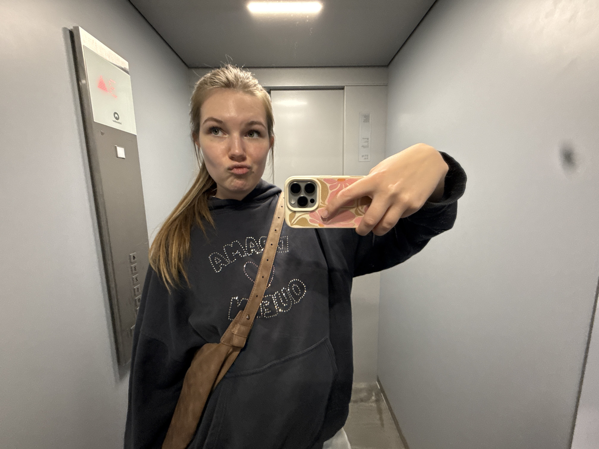 A young woman is taking a selfie in what appears to be an elevator. She is wearing a dark hoodie with rhinestones, a brown shoulder bag, and has her blonde hair partially pulled back. Her phone has a floral case. The elevator has a grey panel displaying an upward arrow, and the walls are a light grey. The location is likely in an urban environment within Vozdovac Urban Municipality, Serbia.